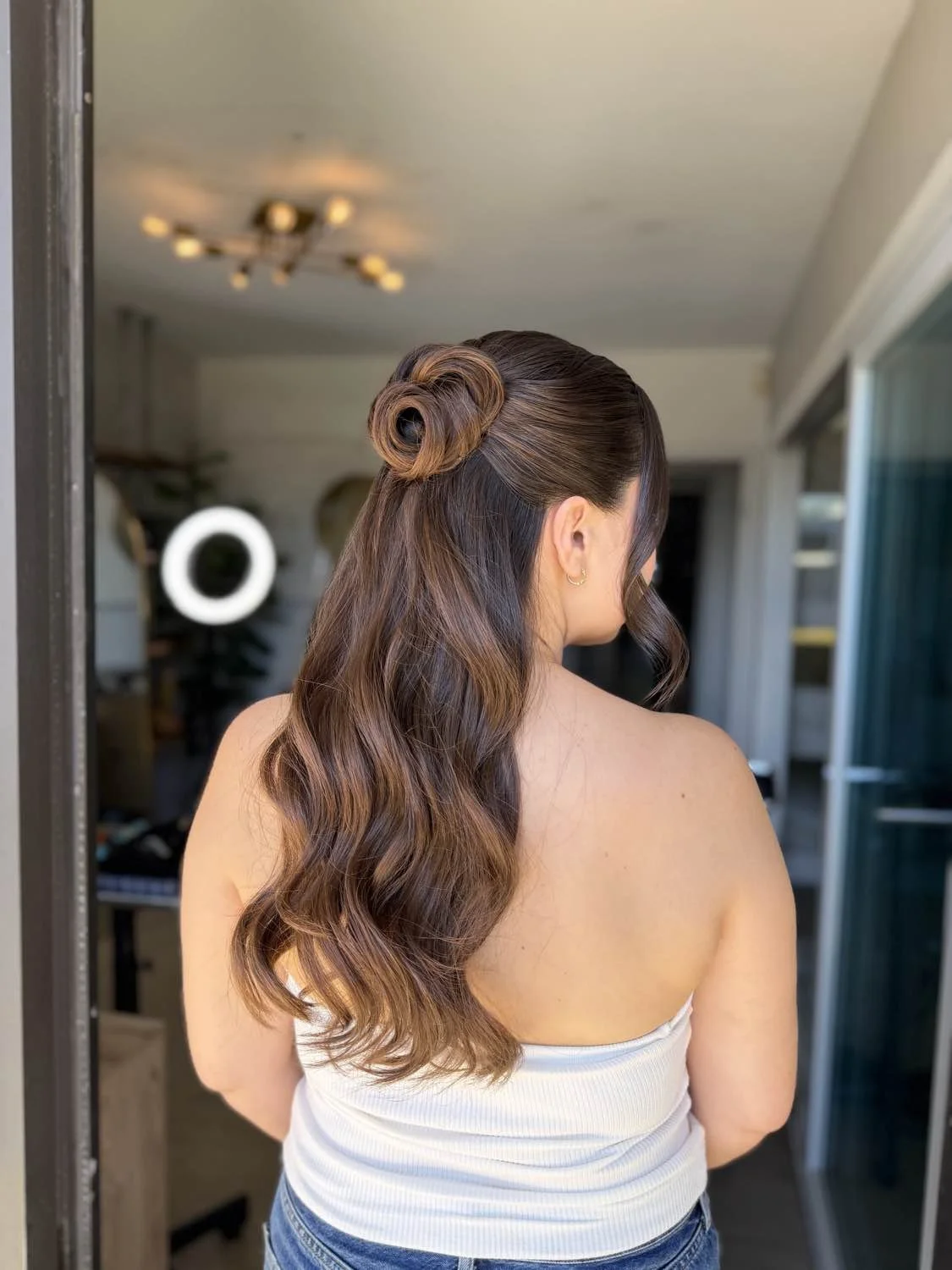 Back view of a woman with long, wavy hair styled in a vintage roll bun on top, standing in a room with a mirror and ceiling light fixture.