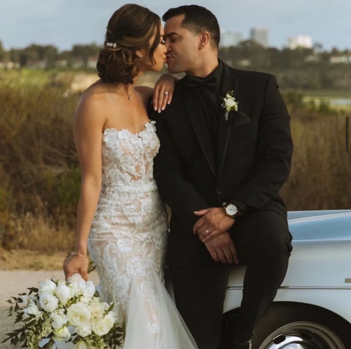 A bride and groom sharing a kiss outdoors on their wedding day. The bride wears a lace wedding dress and holds a bouquet of white flowers. The groom is dressed in a black suit with a boutonniere, sitting on a vintage car.