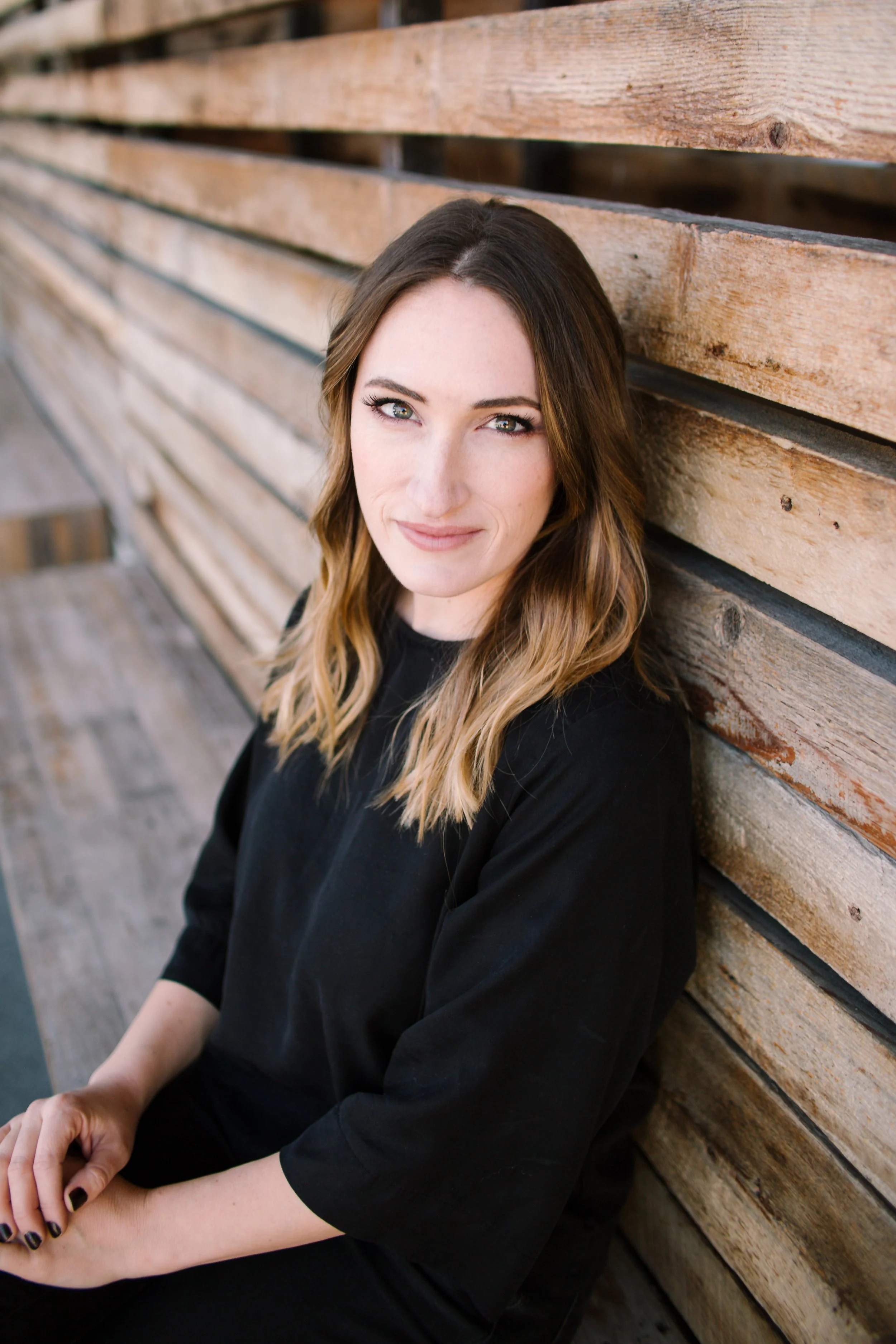 A woman with long brown hair and blue eyes sitting against a wooden wall, wearing a black top.