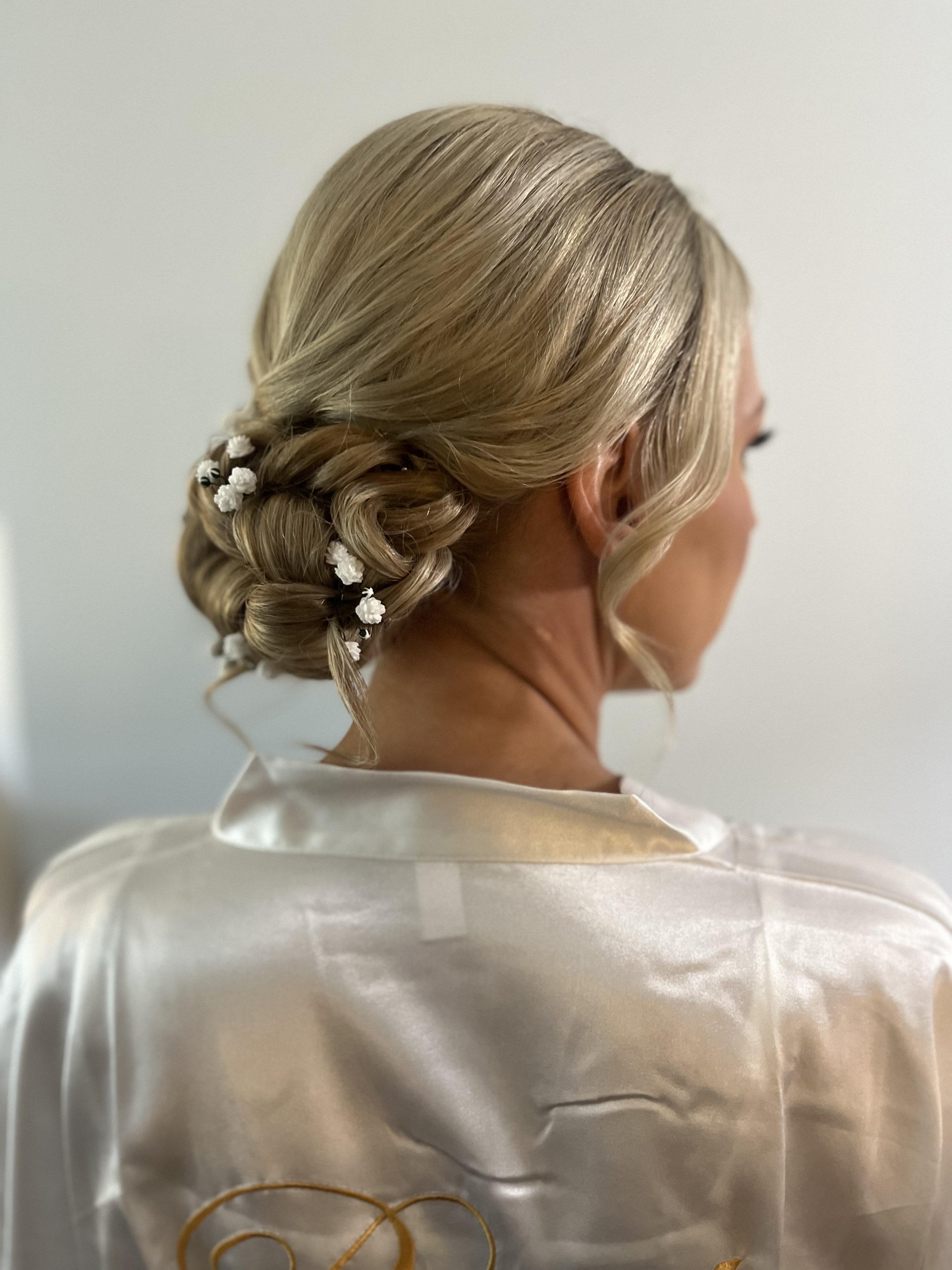 A woman with blonde hair styled in an elegant updo with curls and small white floral decorations, wearing a satin or silk white garment.