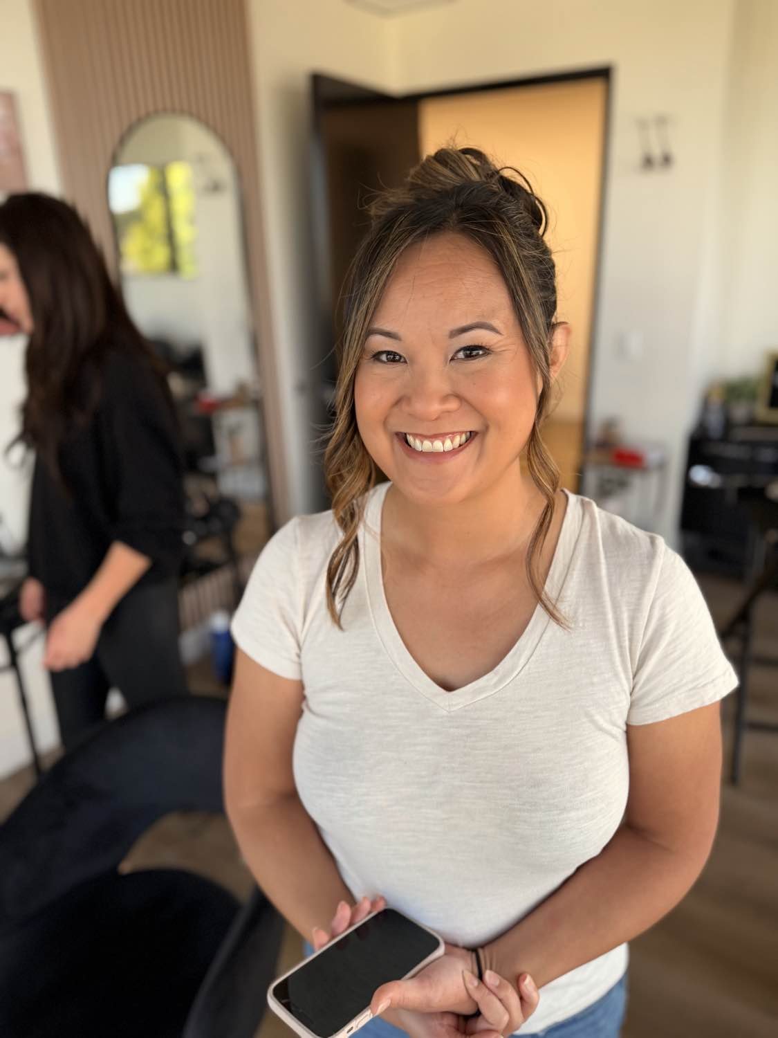 A woman smiling, holding a smartphone, in a room with a mirror and office supplies, with another woman in black in the background.