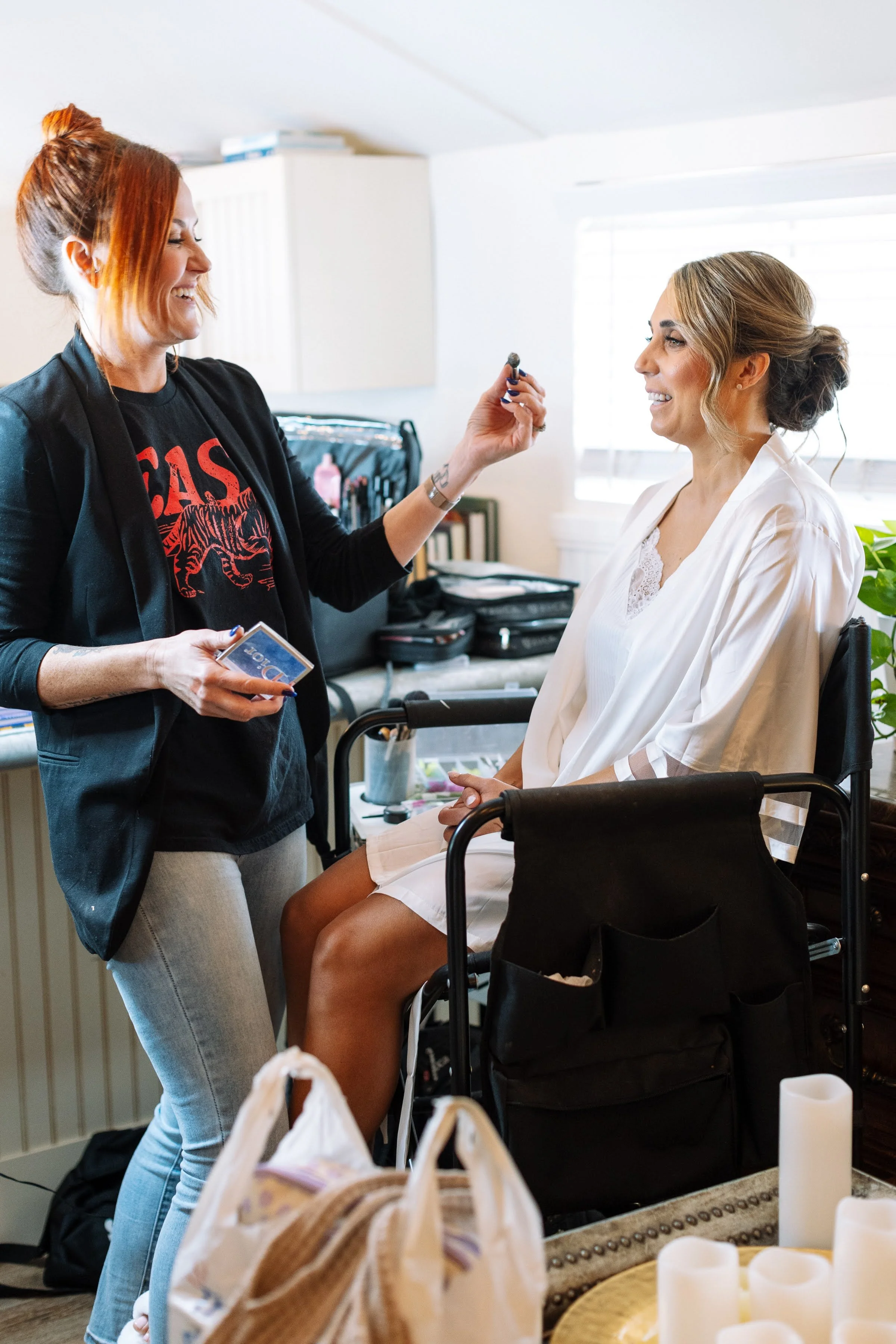 A woman with reddish hair and a woman with blonde hair are smiling at each other in a room with makeup and beauty products. The woman with blonde hair is sitting in a wheelchair wearing a white robe, while the other woman is standing and wearing a black jacket and a graphic T-shirt.