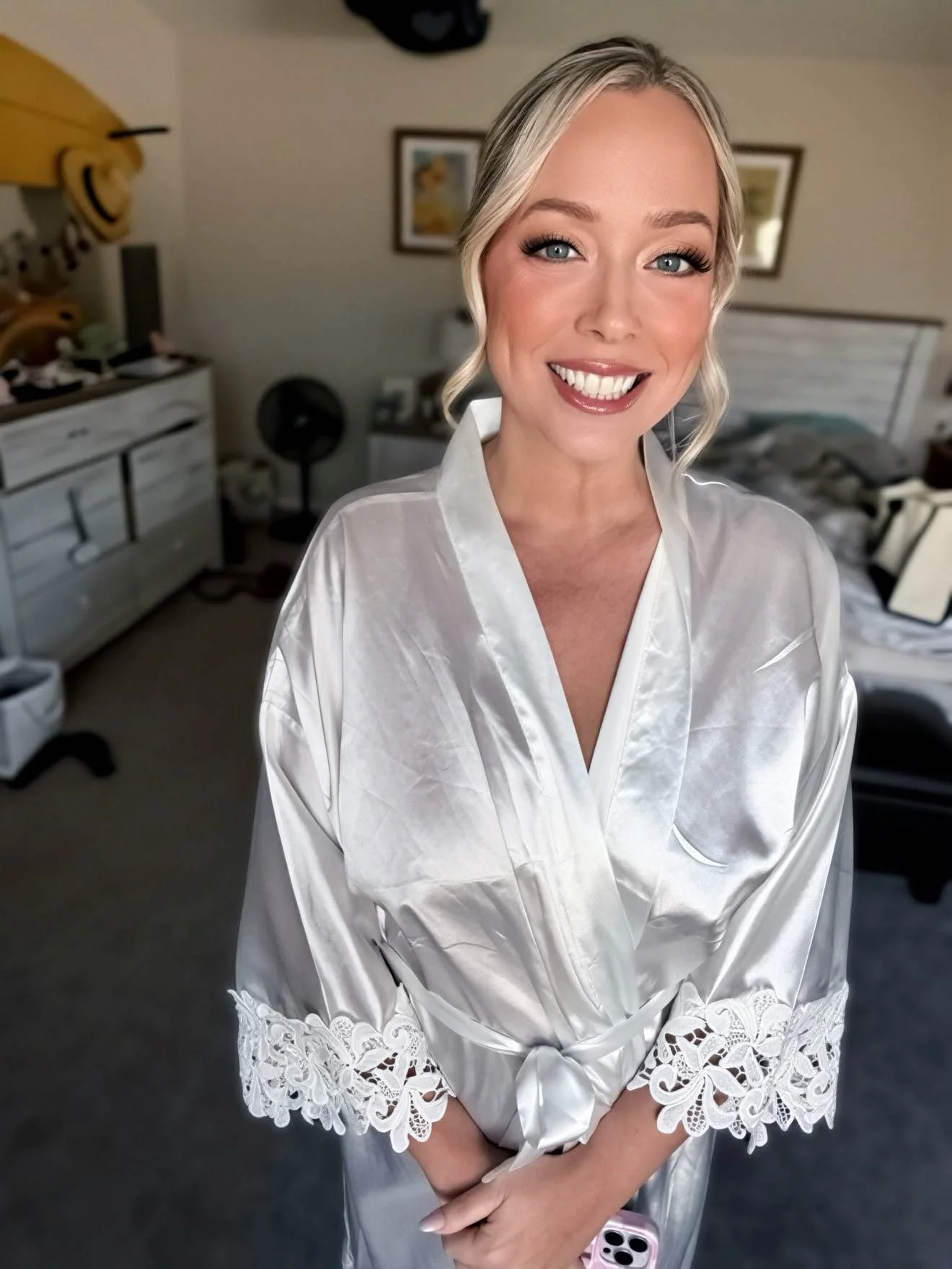 A woman with blonde hair styled in loose waves smiling, wearing a white satin bathrobe with lace details on the sleeves.