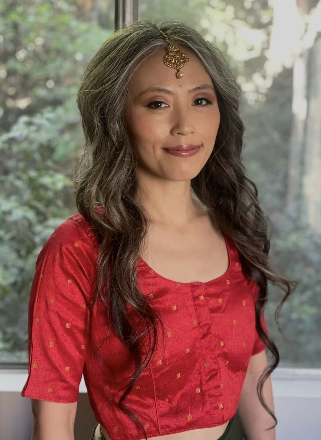 A woman with long, wavy gray hair wearing a red traditional Indian outfit and a decorative headpiece, standing indoors near a window with a greenery background.