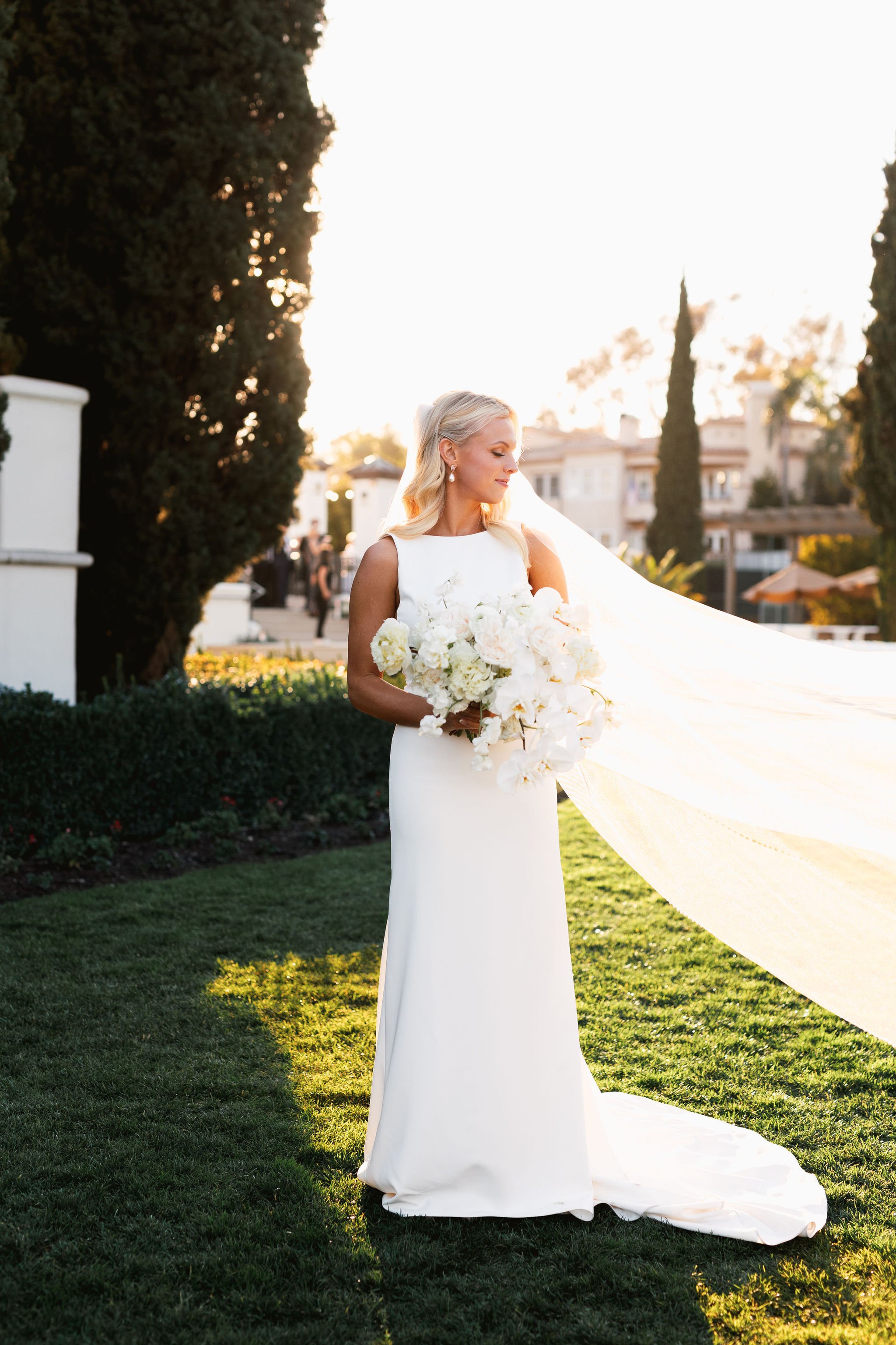 A bride in a white wedding dress holding a bouquet of white flowers standing outdoors during sunset with trees and buildings in the background.