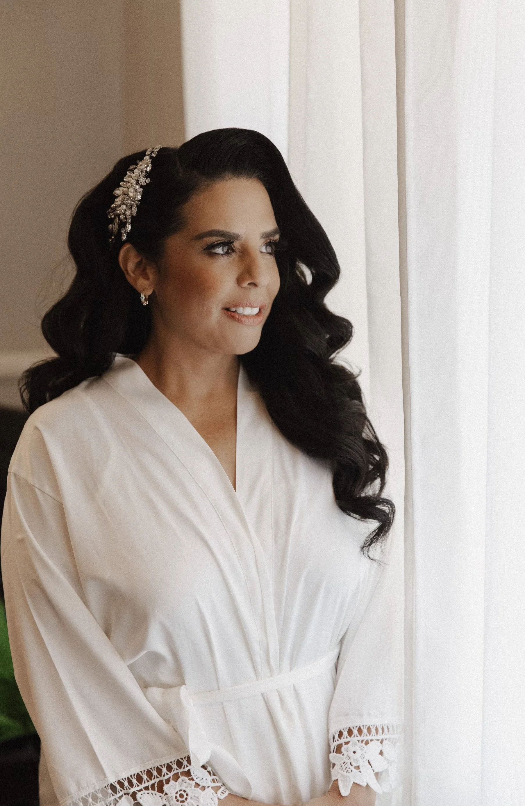 A woman with dark, wavy hair styled in vintage waves, wearing a pearl and rhinestone hair accessory and a white robe with lace-trimmed sleeves, standing by a window with white curtains.
