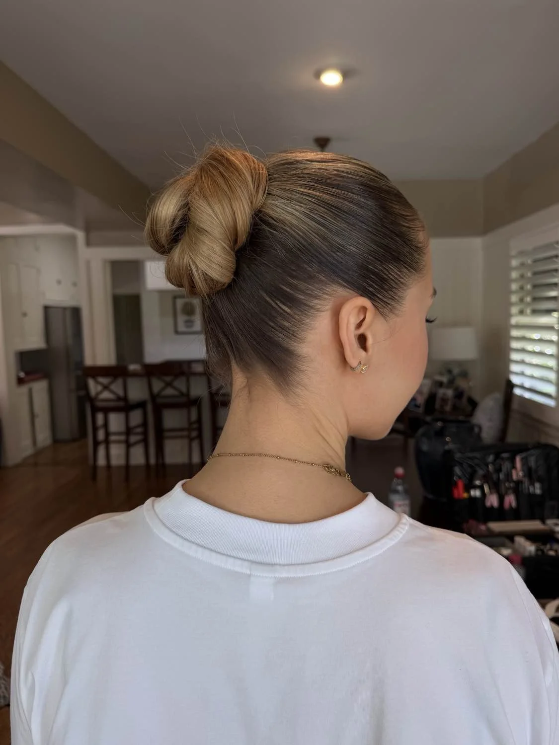 A woman with her hair styled in a sleek, high bun with gradient coloring, wearing a white shirt, gold earrings, a necklace, and standing inside a house with a kitchen area in the background.