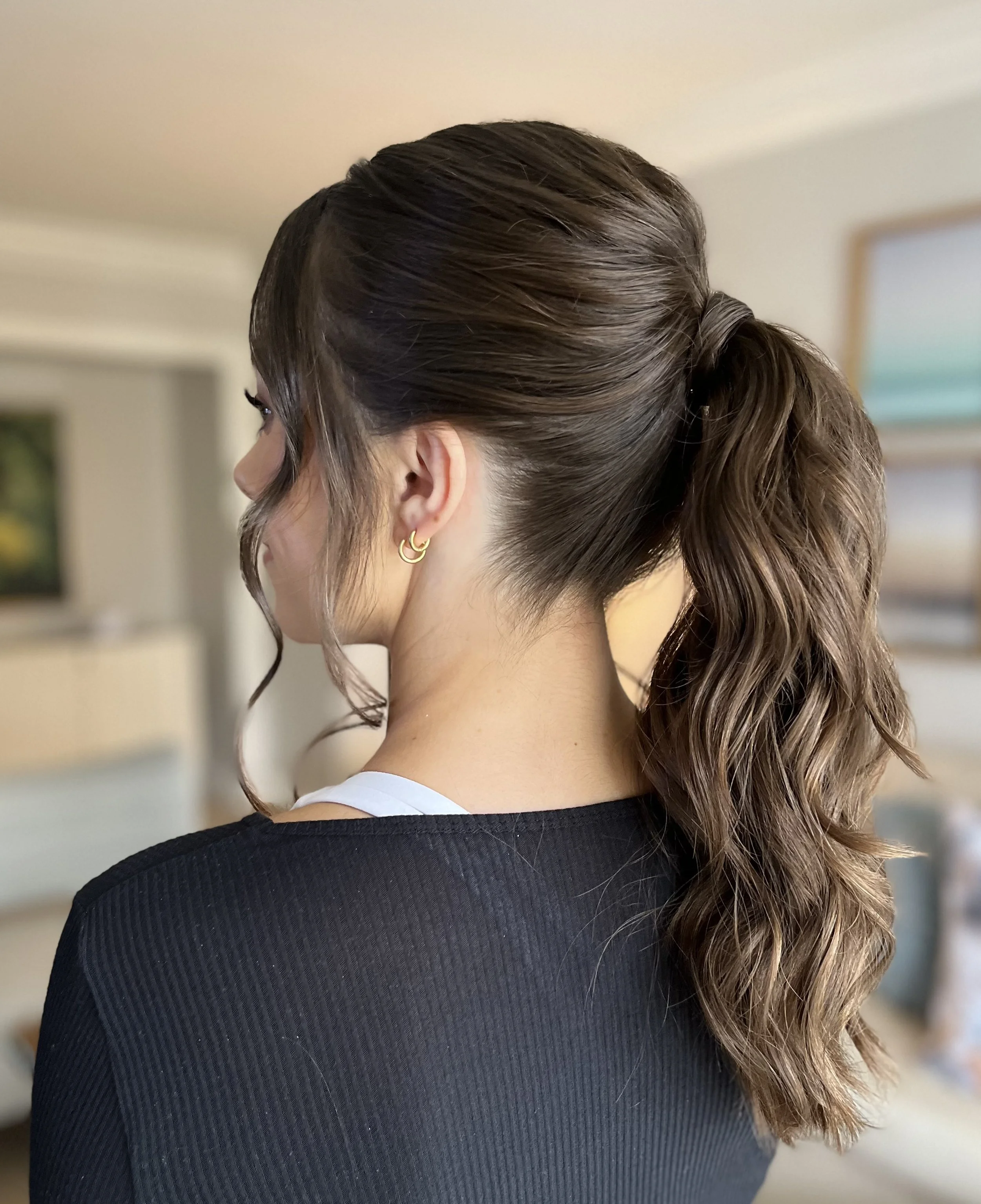 Side view of a woman with brown, wavy hair styled in a half-up half-down ponytail, wearing gold hoop earrings and a black top.
