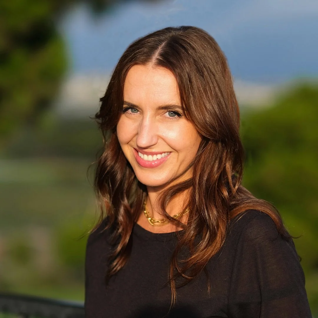 A woman with long brown hair smiling outdoors, with a green landscape and blue sky in the background.