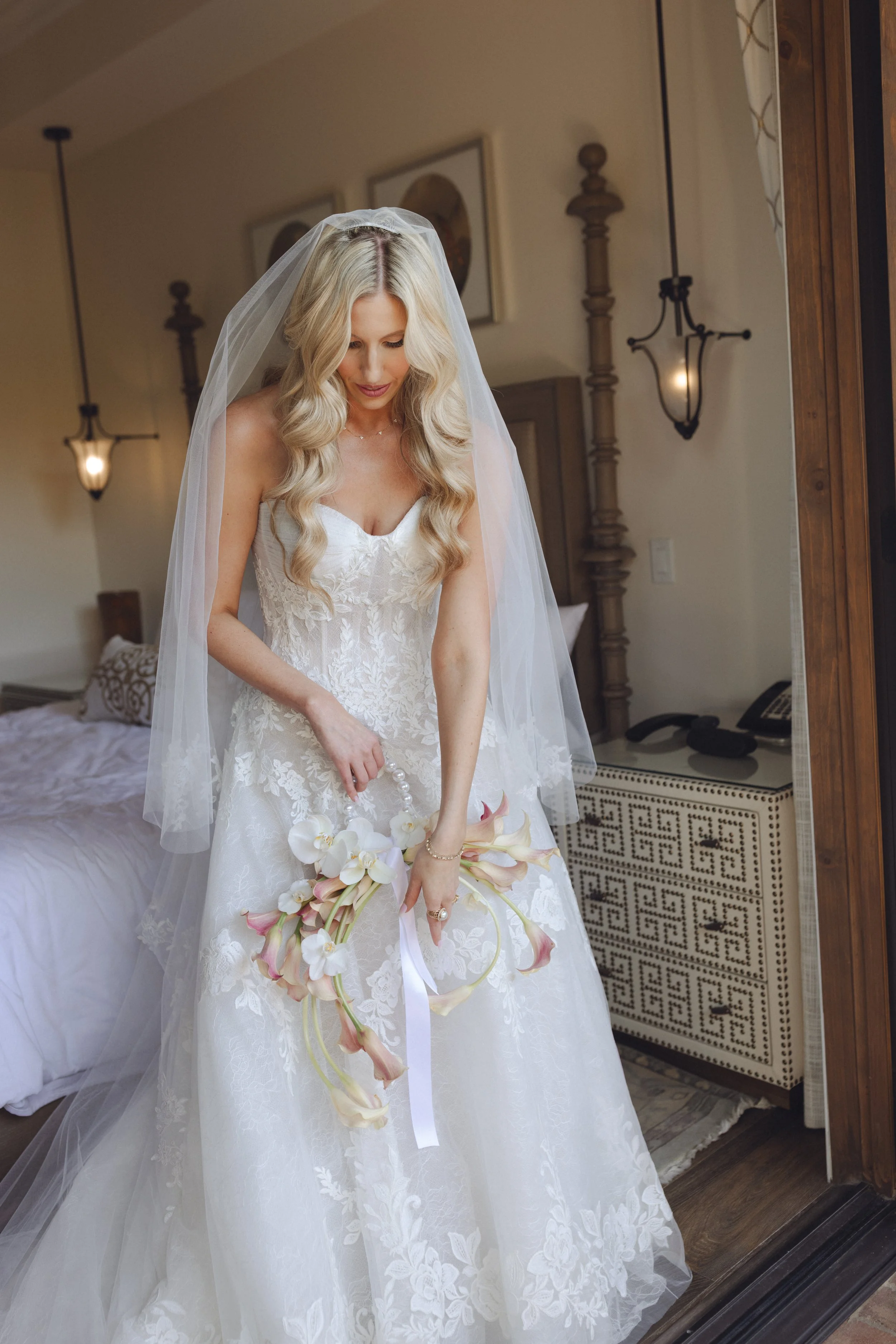 A bride in a white wedding dress with lace details, long wavy blonde hair, and a veil, holding a bouquet of orchids and calla lilies, standing in front of a mirror in a bedroom with warm lighting and wooden furniture.