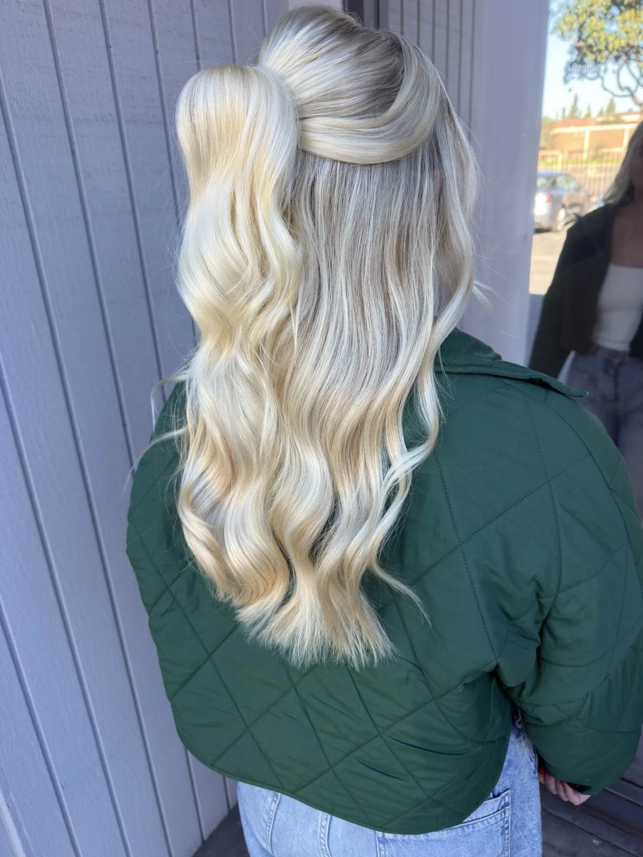 Back view of a woman with long, wavy, blonde hair wearing a dark green quilted jacket and light blue jeans, standing outdoors near a gray wooden wall.