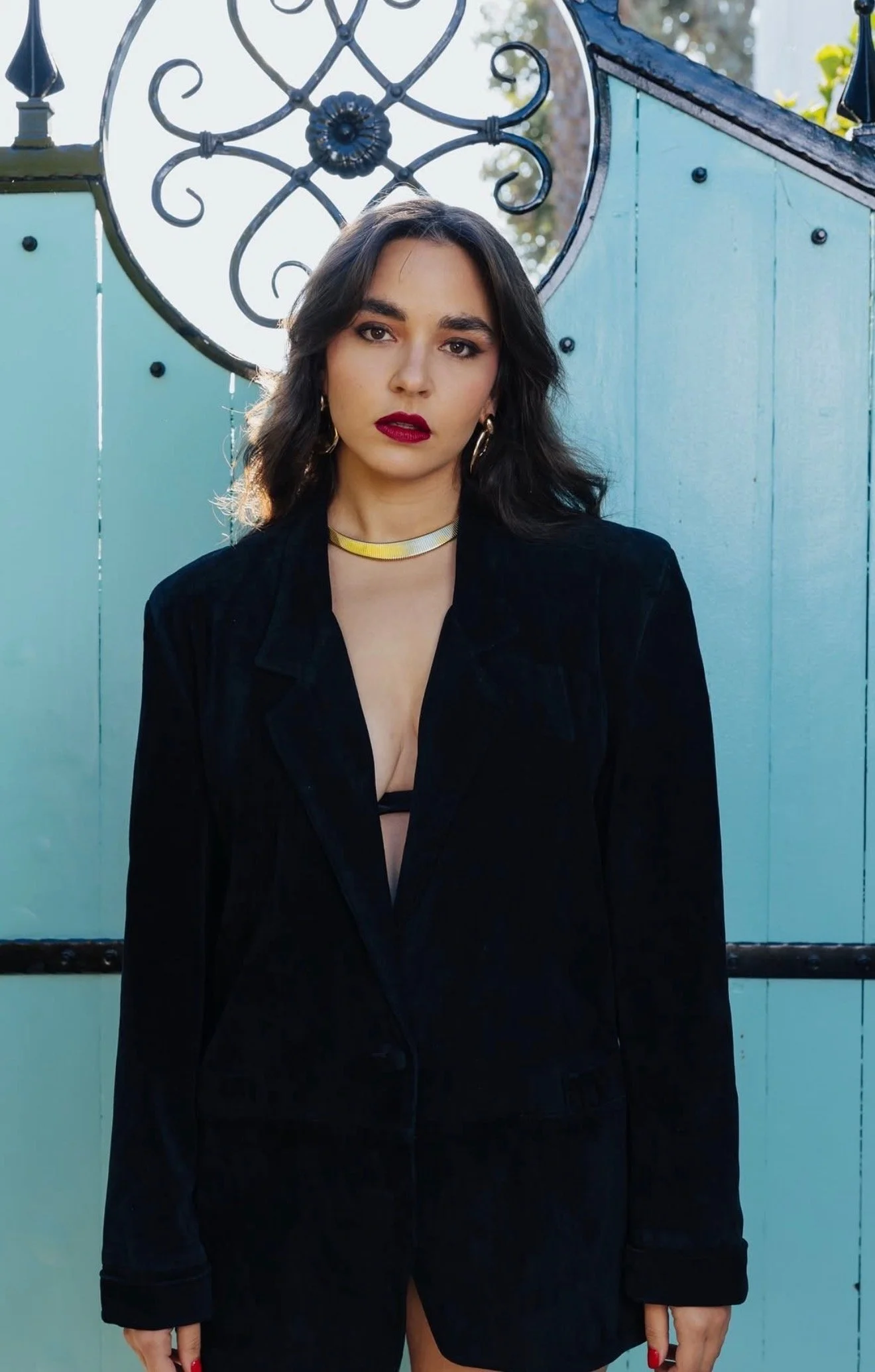 A woman with dark wavy hair wearing a black blazer and bold red lipstick standing in front of a light blue wooden fence with a decorative wrought iron circle above her head.