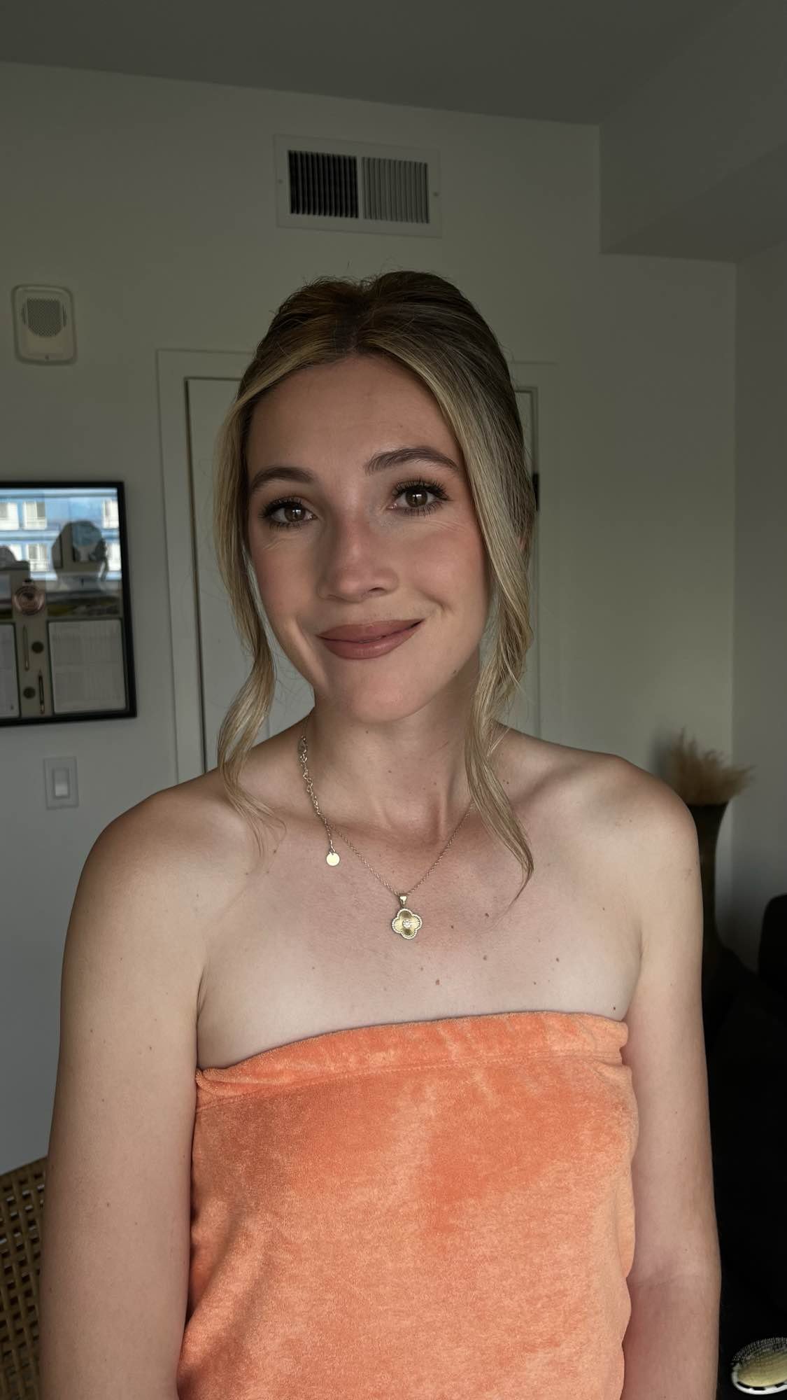 A woman with blonde hair and light skin smiling, wearing an orange strapless top and a gold necklace with a four-leaf clover pendant, in an indoor setting.
