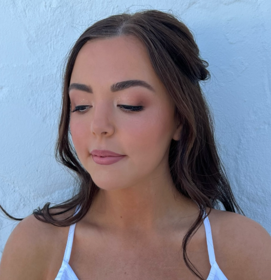 Close-up of a young woman with long brown hair and makeup, wearing a white top, against a light blue wall.