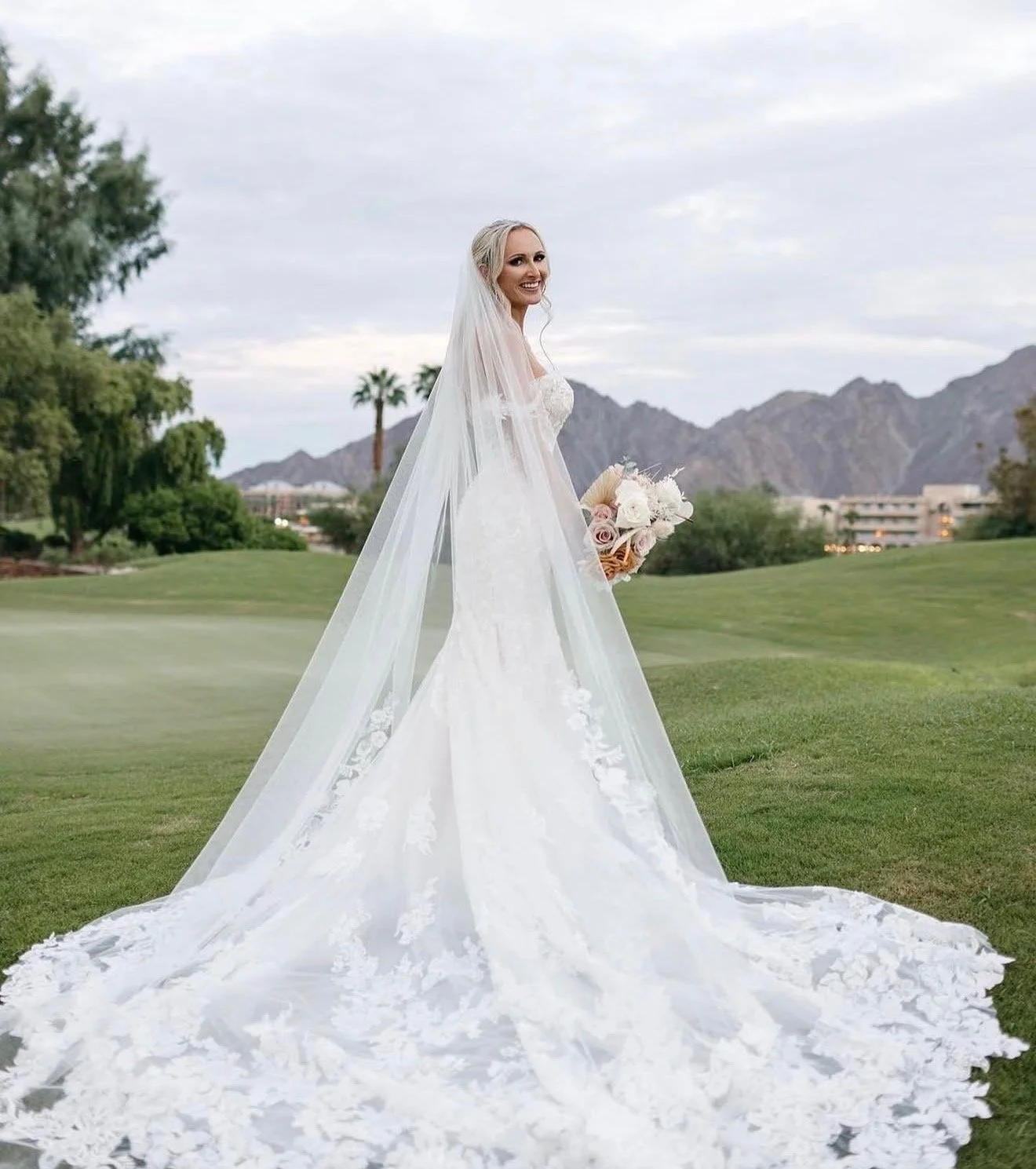 Bride in a white wedding gown holding a bouquet outdoors with mountains, trees, and cloudy sky in the background.