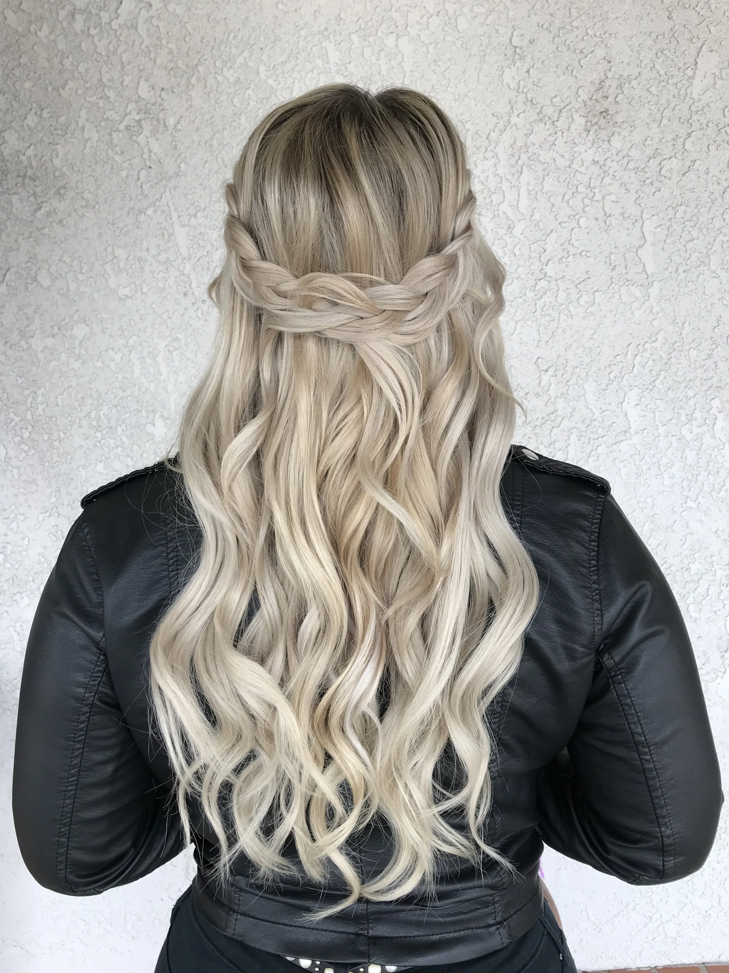 Back view of a woman with long, wavy, blonde hair wearing a black leather jacket and standing against a textured white wall. Her hair features a crown braid encircling her head.