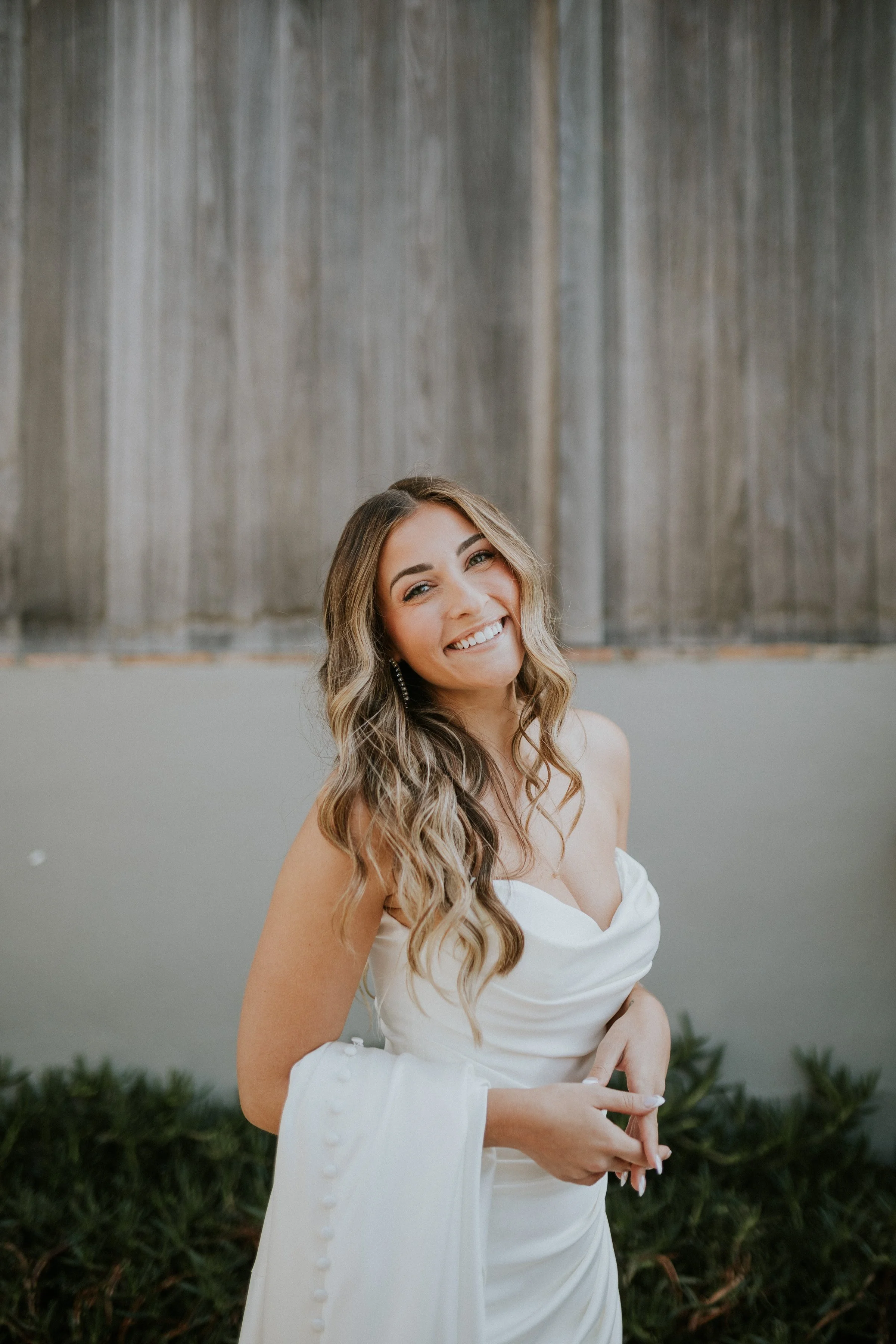 A smiling woman with wavy hair wearing a white off-shoulder dress, standing outdoors in front of a gray wall with green plants at the base.
