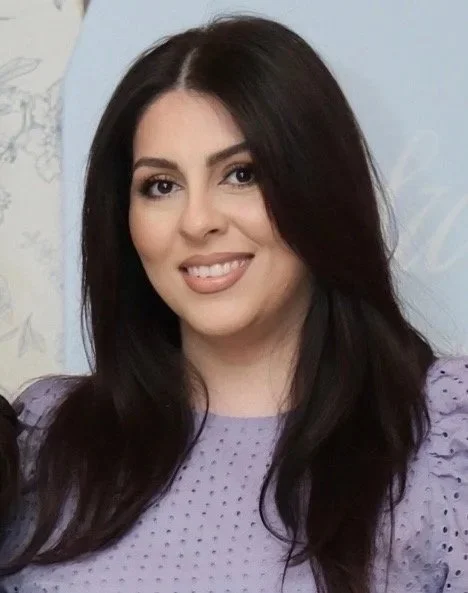 A woman with long dark hair, smiling in a purple polka dot top, posing indoors against a light-colored background.
