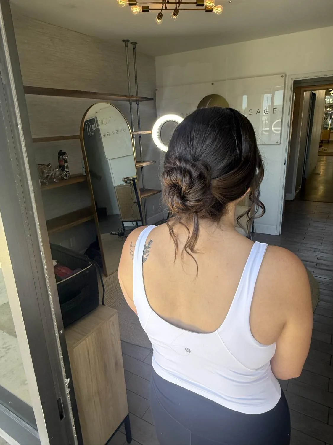 A woman with dark hair styled in an elegant low bun with loose tendrils, wearing a white tank top, is seen from behind in a room with modern decor and a mirror reflecting her back.