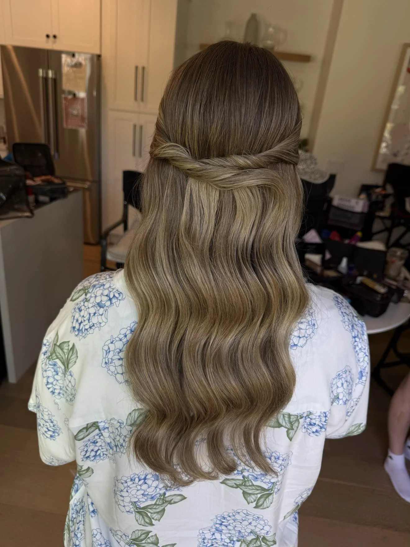 Back of a woman with long, styled blonde hair that has loose waves and a twisted braid detail at the crown, wearing a white dress with blue floral patterns.