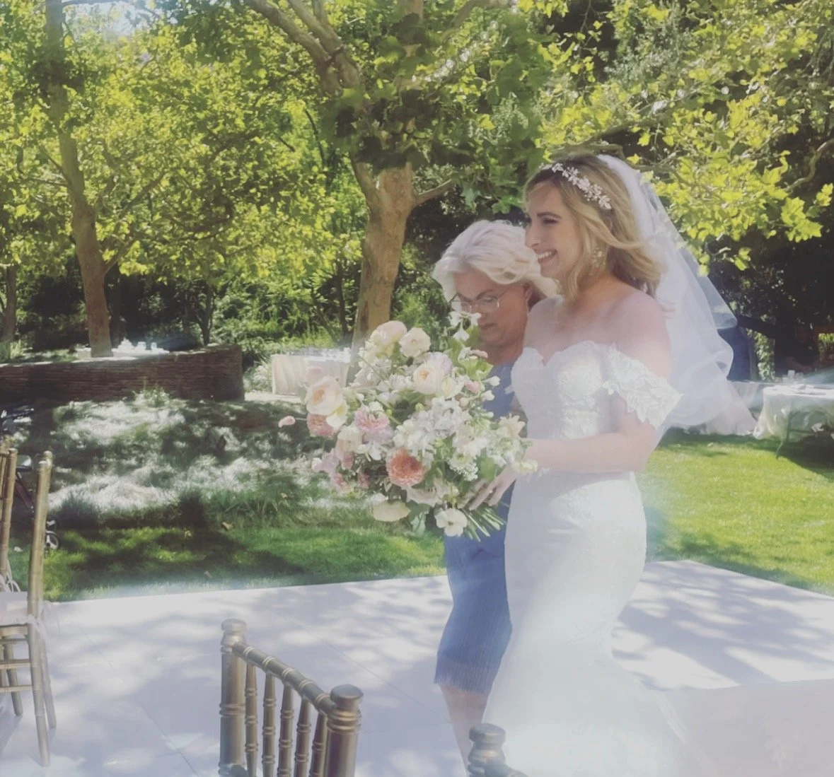 A bride in a white wedding dress and veil holding a large bouquet of flowers, standing outdoors with a woman, surrounded by green trees and grass.