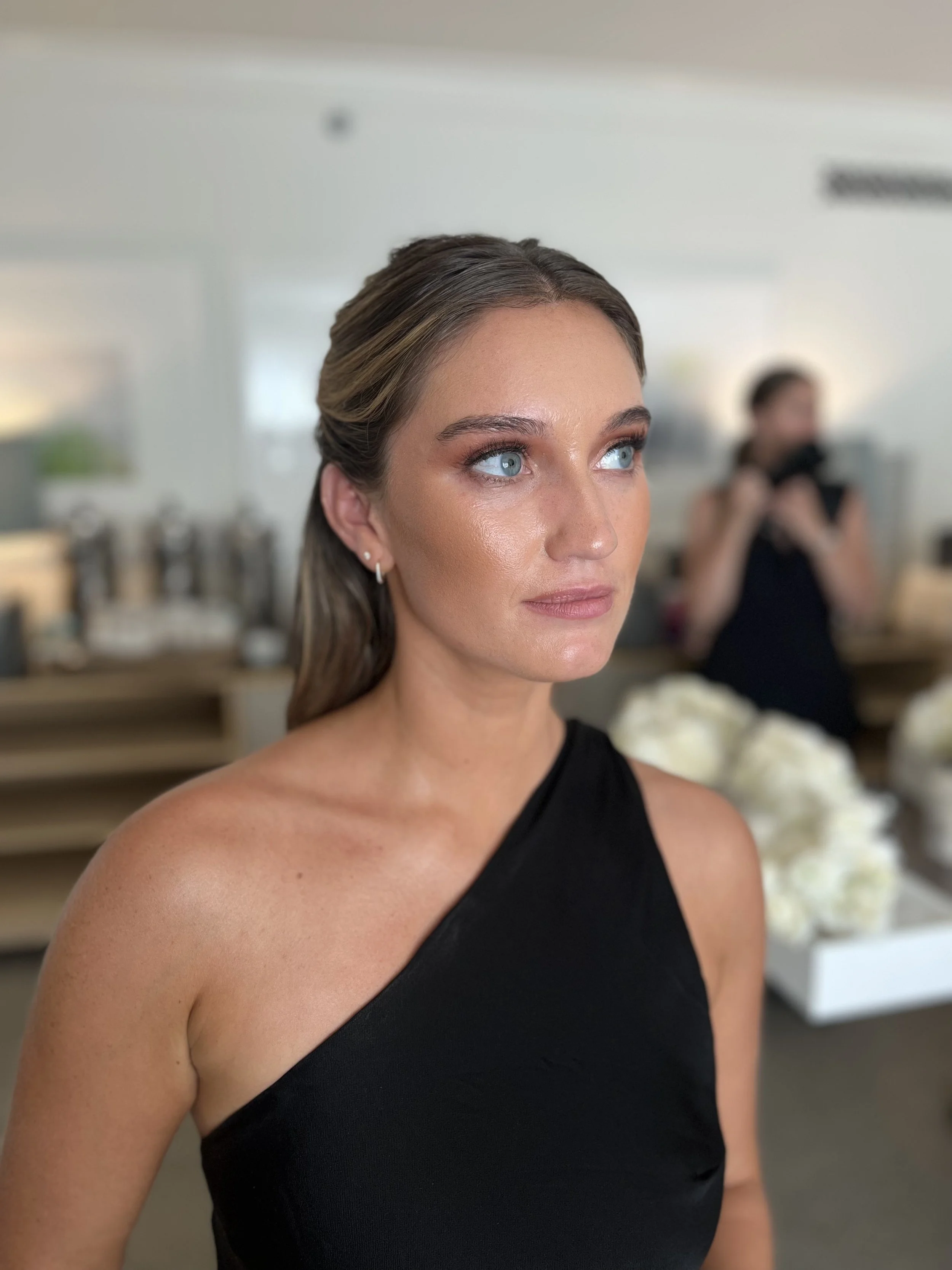 Close-up of a woman with blue eyes and light brown hair styled back, wearing a black sleeveless top, in an indoor setting with blurred background.
