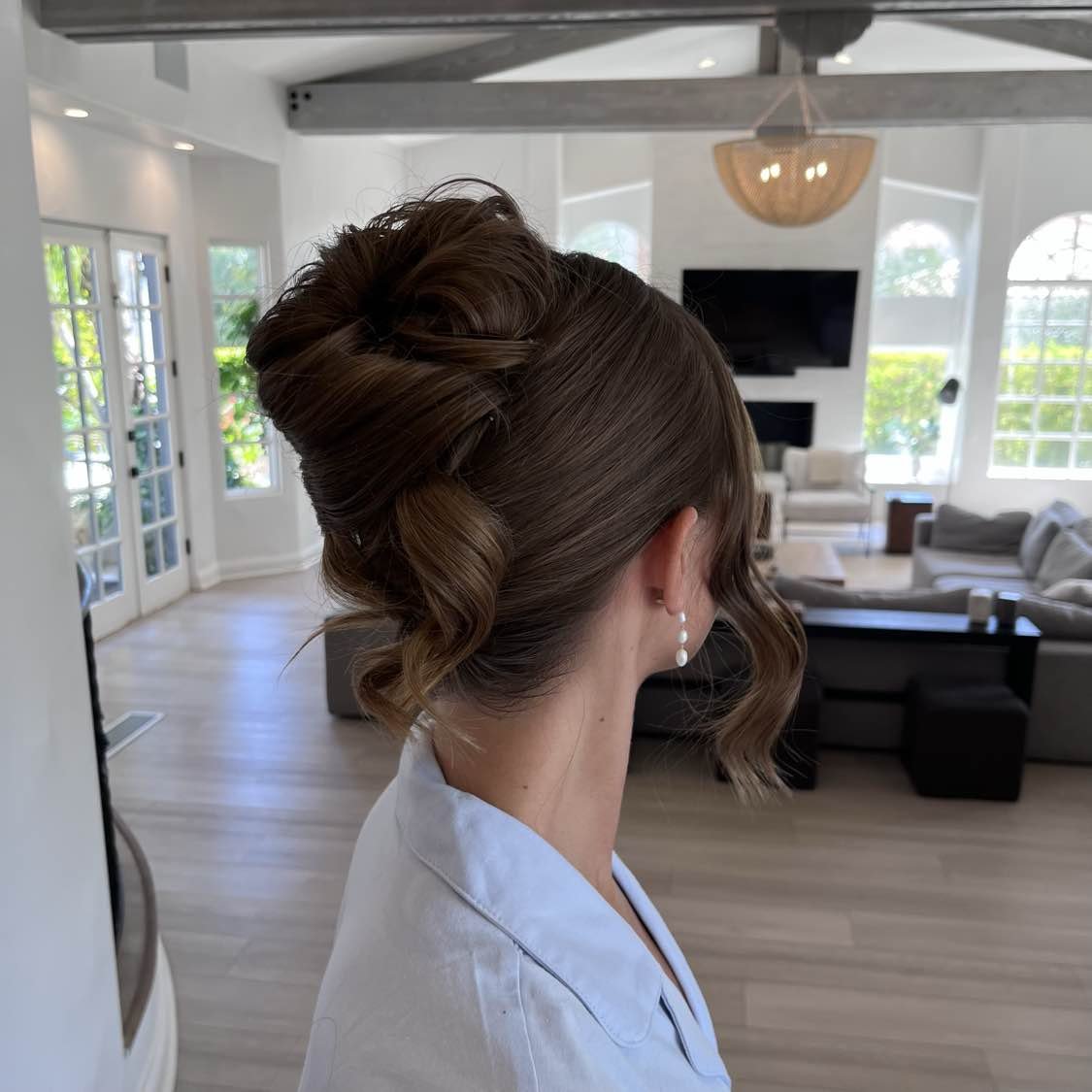 A woman with styled brown hair in an elegant updo with loose curls, wearing pearl earrings, in a bright, modern living room with large windows and contemporary furniture.