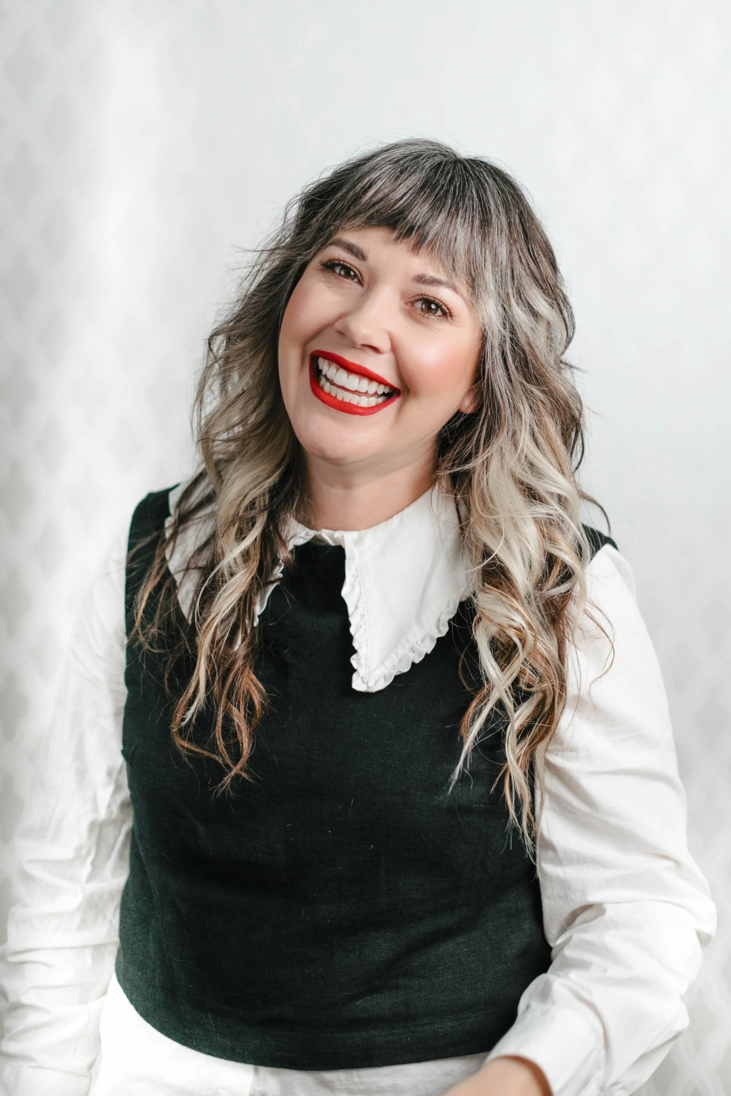 A woman with long wavy hair and bangs, wearing a white shirt with a black vest, smiling with red lipstick against a plain white background.