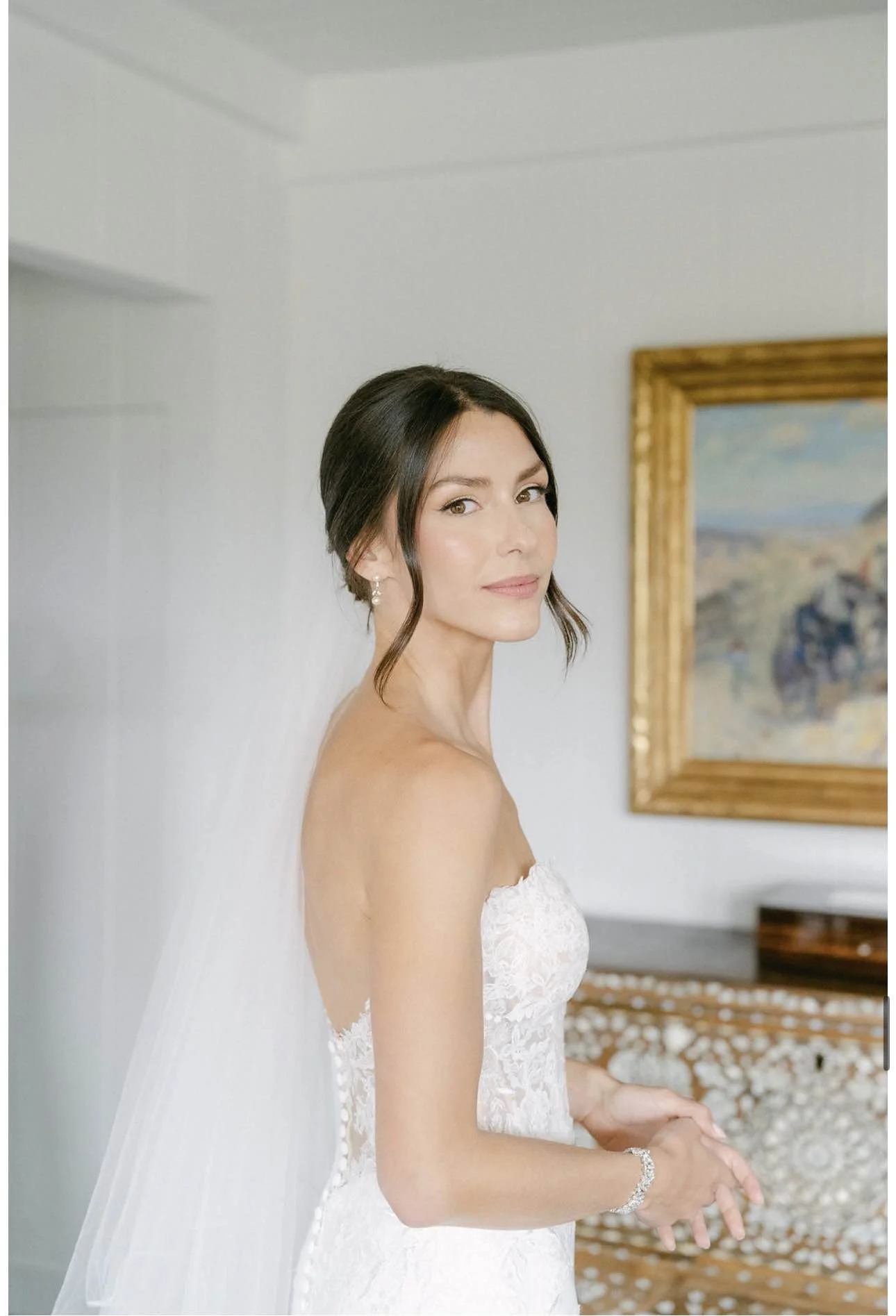 A woman in a wedding dress standing indoors with a portrait painting in the background.