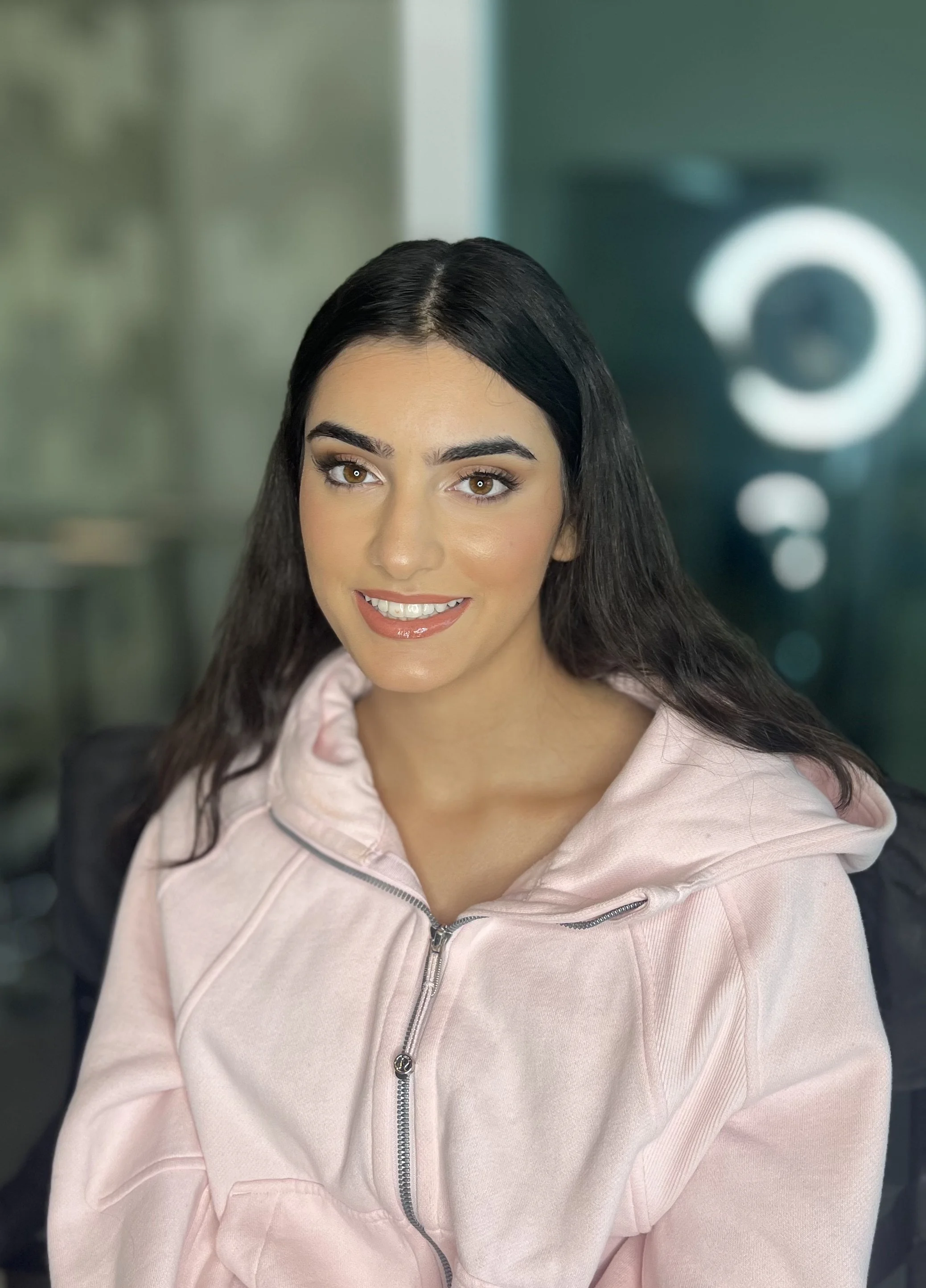 A woman with long dark hair, brown eyes, and light makeup, smiling and wearing a light pink zip-up hoodie.