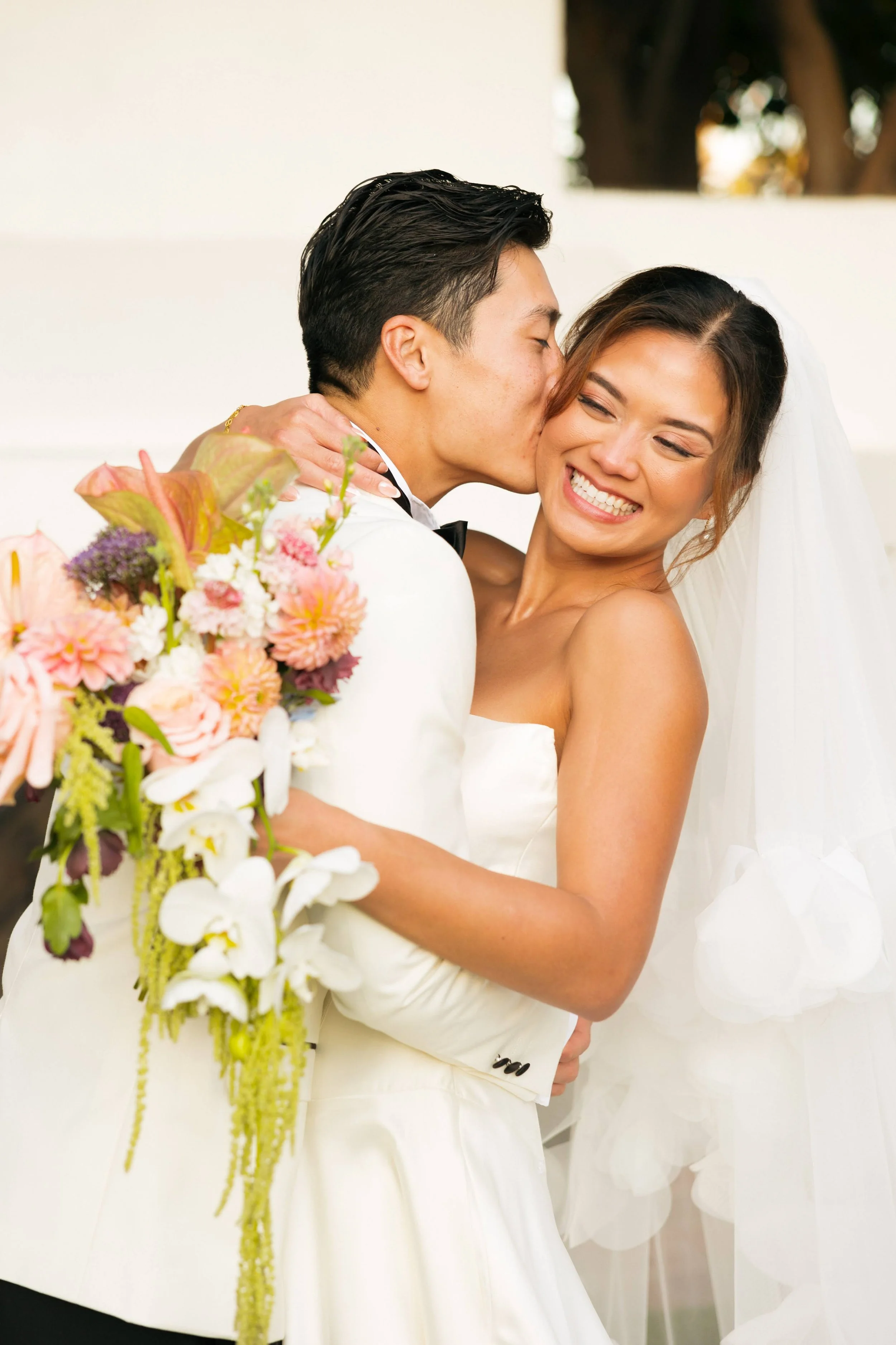 A newlywed couple embracing, with the groom giving a kiss on the cheek to the bride, who is smiling with closed eyes, holding a bouquet of flowers.