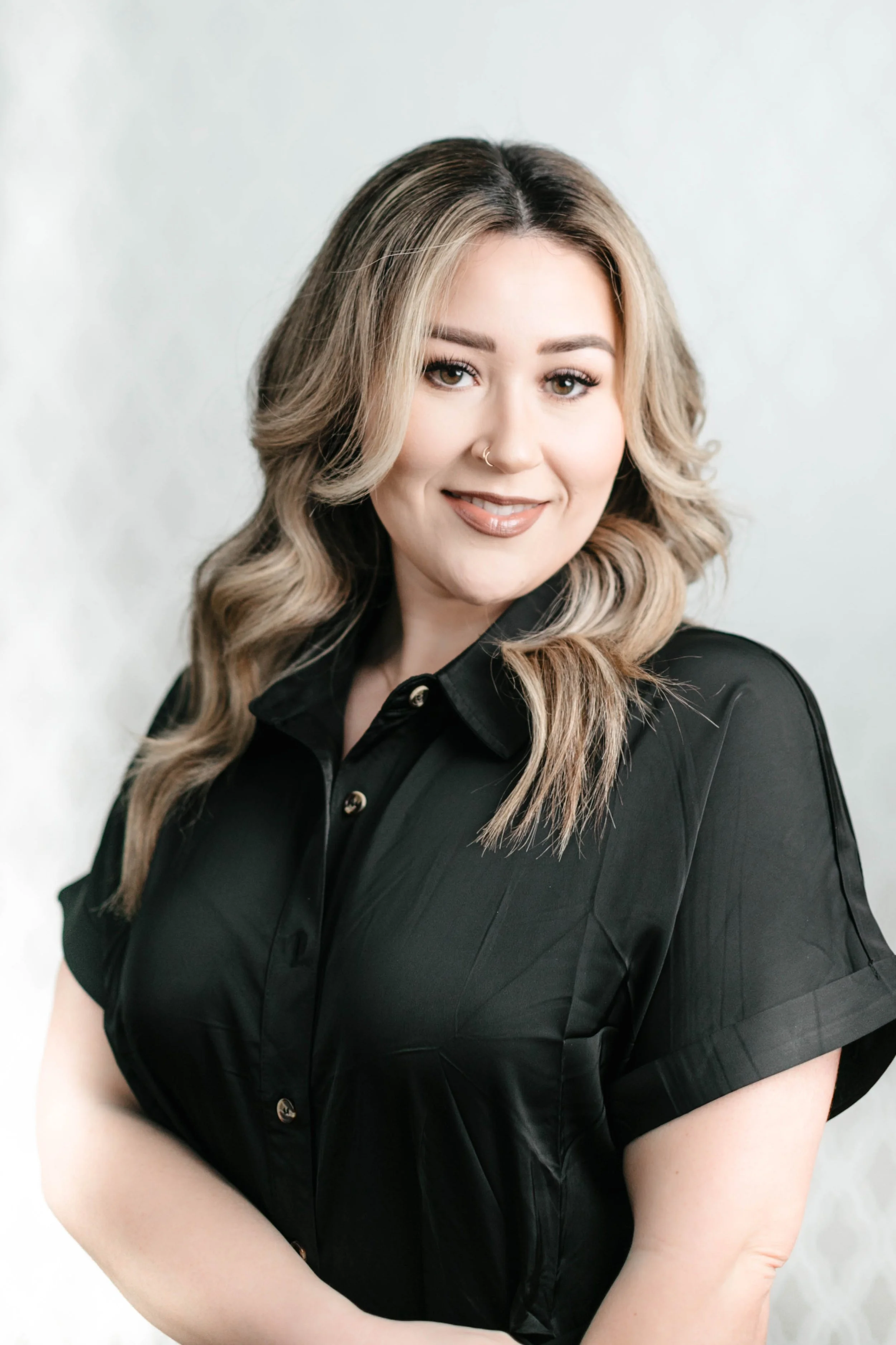 A woman with long, wavy blonde hair, light skin, and a nose ring, wearing a black shirt, smiling softly, against a plain light-gray or white background.