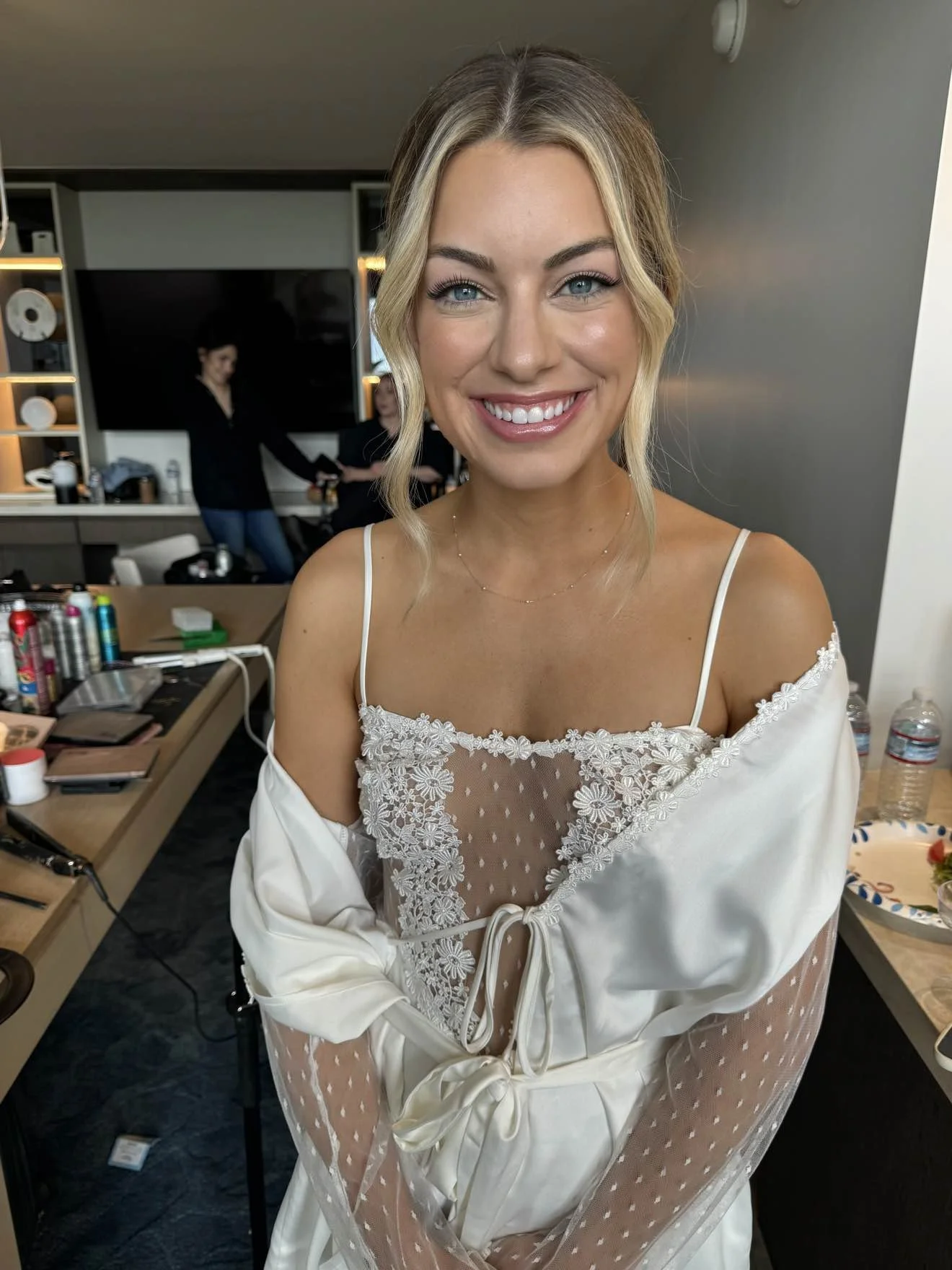 A smiling woman with blonde hair and blue eyes in a white lace top in a room with makeup and hairstyling supplies, with two people in the background.