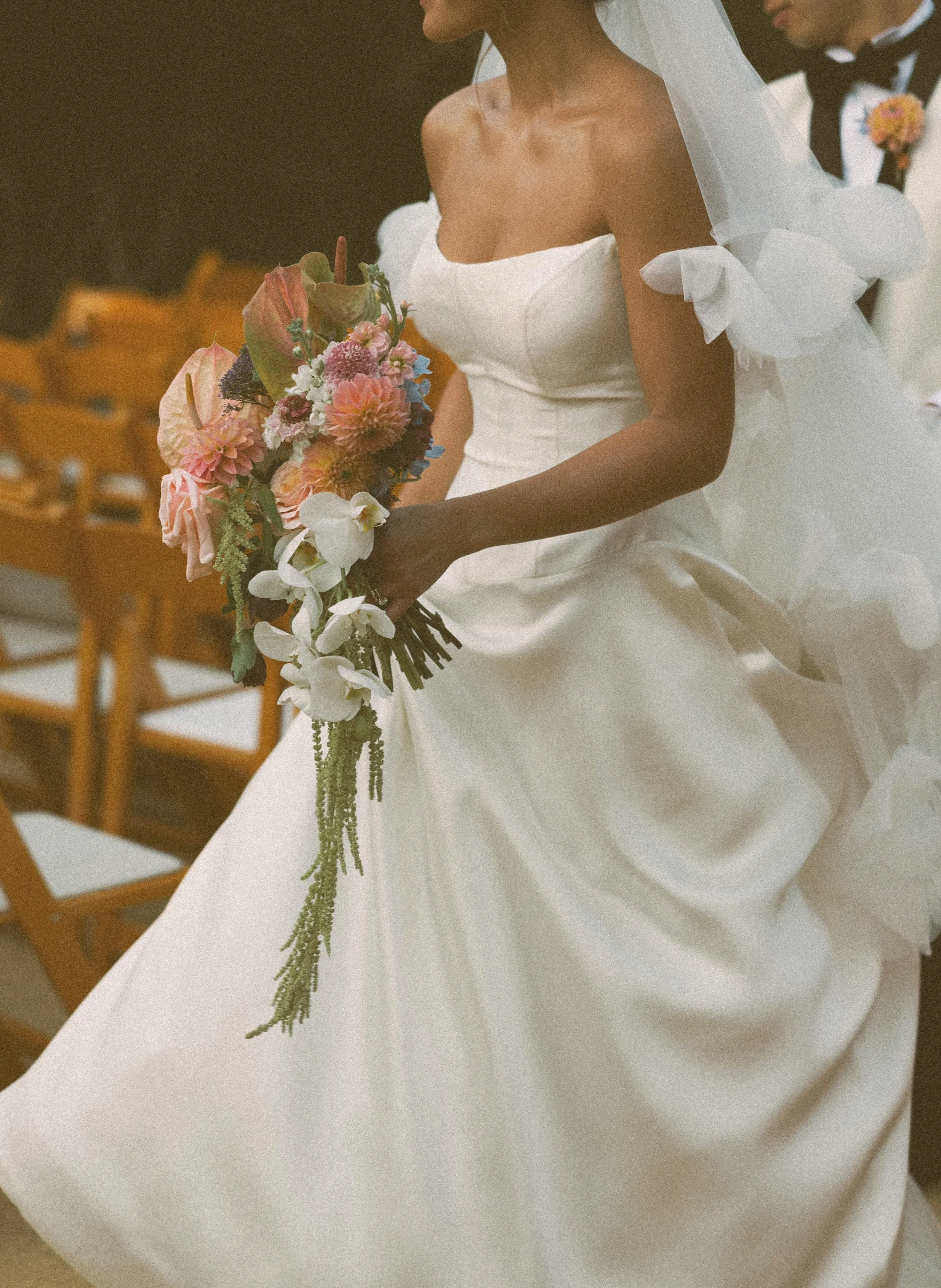 A bride in a white strapless wedding dress holding a bouquet of pink, white, purple, and green flowers, with a veil on her head, at an indoor wedding ceremony.