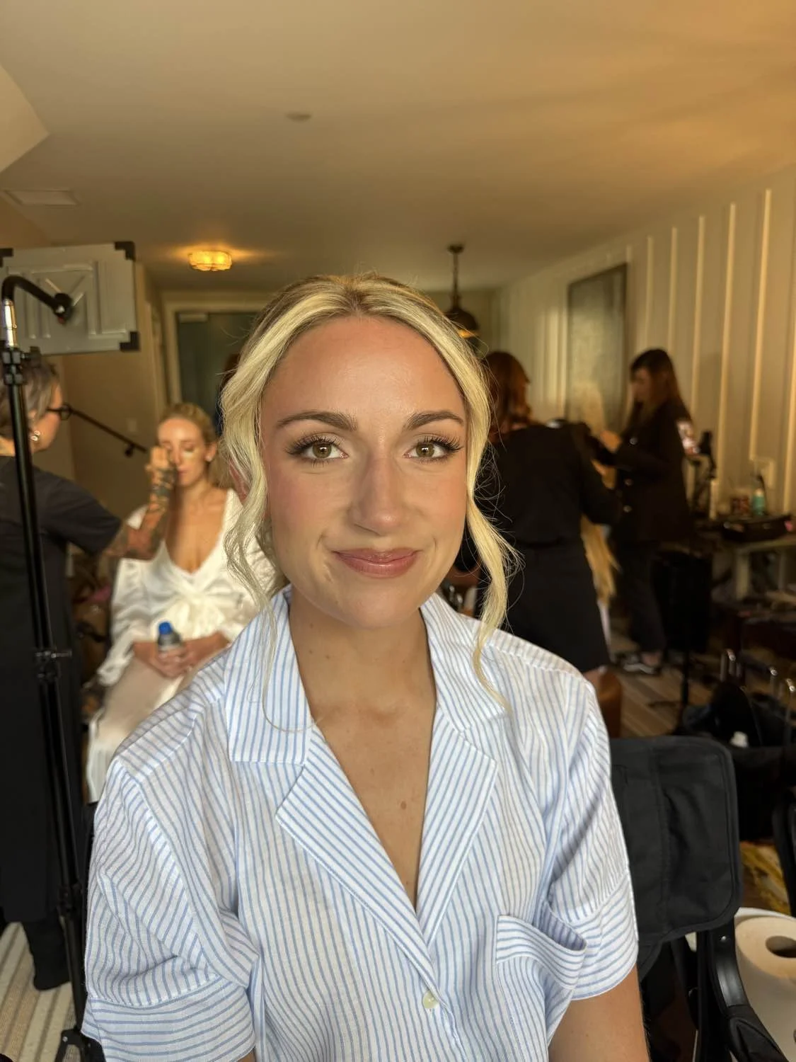 A woman with blonde hair and a blue striped shirt sitting in a room with makeup artists and hairstylists in the background, preparing for a photoshoot or event.