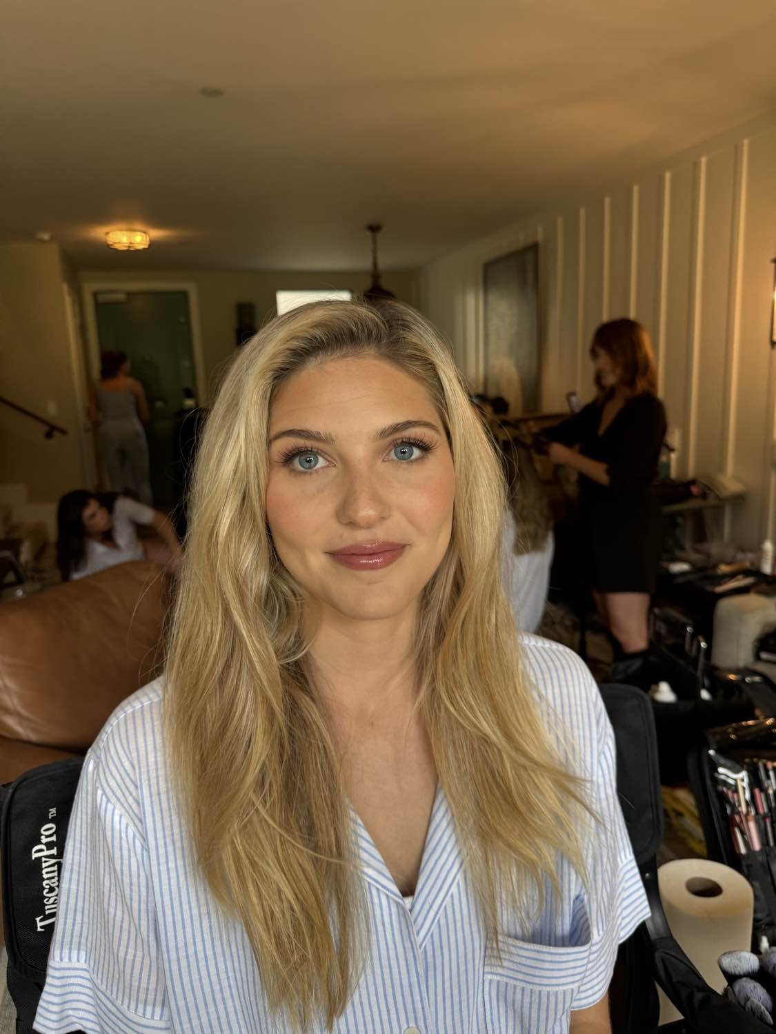 A woman with long blonde hair and blue eyes looking at the camera, in a room with other women and a person working on makeup or hair in the background.