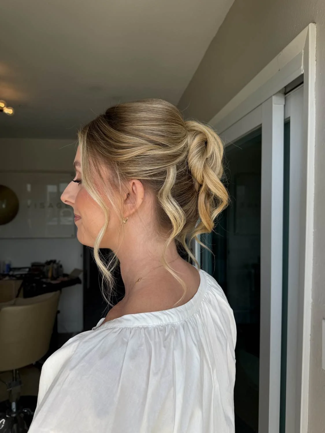 A woman with blonde hair styled in a loose, wavy updo, wearing a white top, stands indoors next to a sliding glass door.