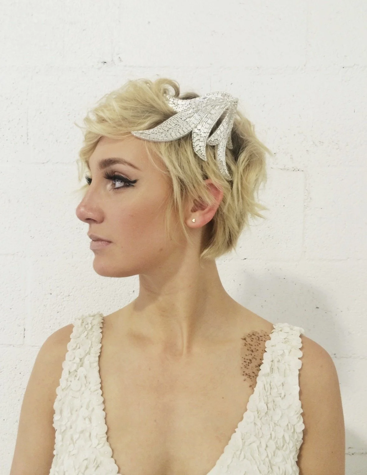 A woman with short blonde hair wearing a decorative silver headband in the shape of a starfish, standing against a white brick wall.