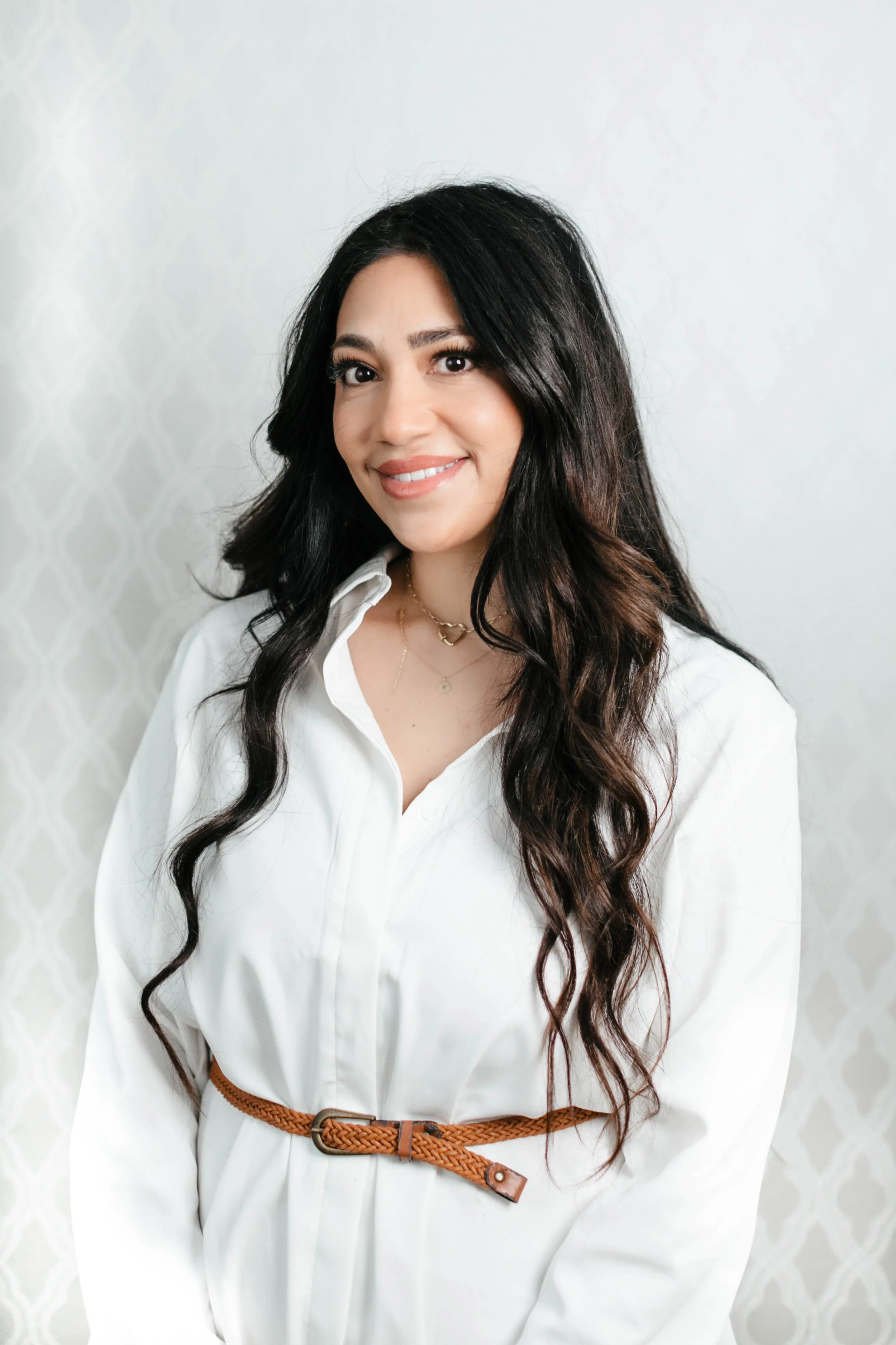 A woman with long dark wavy hair, wearing a white button-up shirt, a brown braided belt, and layered gold necklaces, smiling and standing in front of a light-colored textured background.