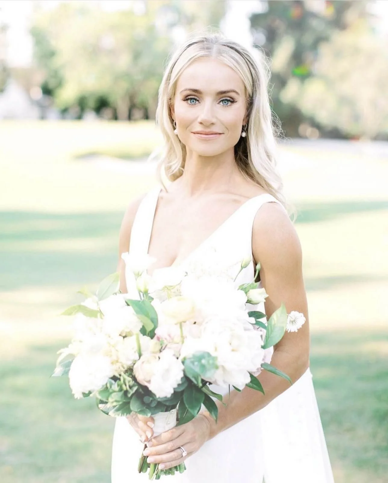 A woman in a white dress holding a large bouquet of white flowers outdoors in a park with green grass and trees in the background.