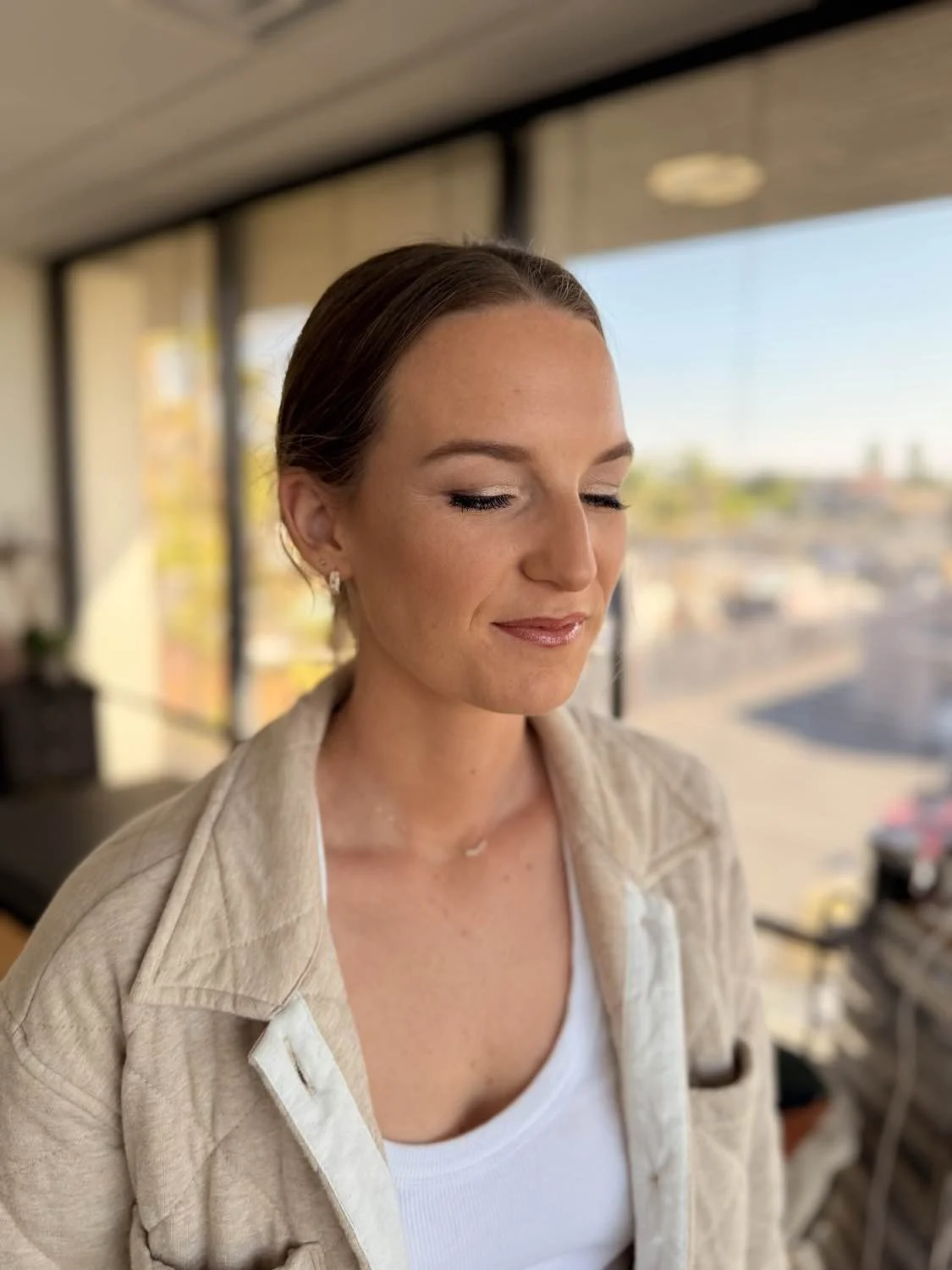 A woman with brown hair and closed eyes, wearing a beige jacket and white top, standing in a room with large windows and cityscape outside.