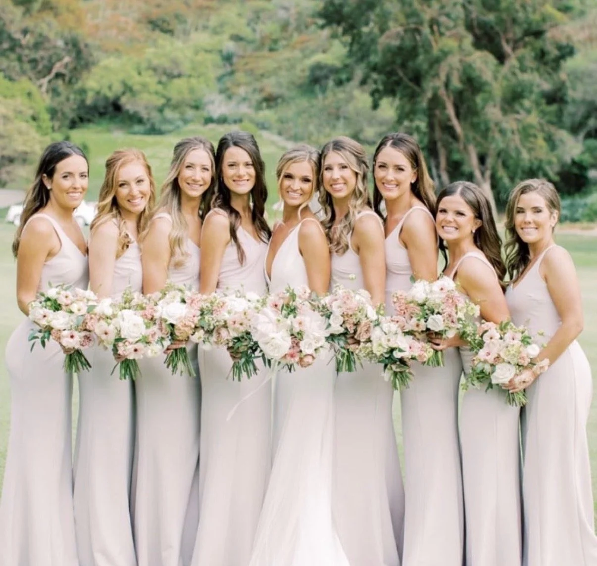 A wedding party of ten women in white dresses holding bouquets of pink and white flowers outdoors.