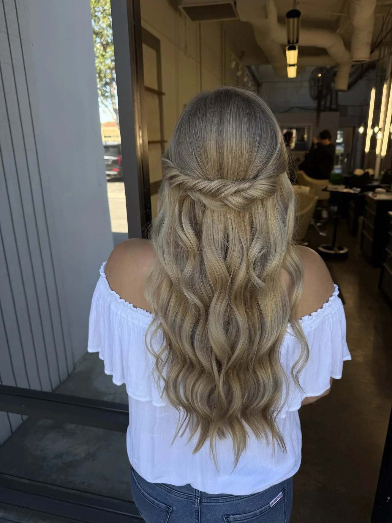 Back view of a woman with long, blonde, wavy hair styled in a half-up twisted crown, wearing an off-shoulder white top and jeans, standing inside a modern salon.