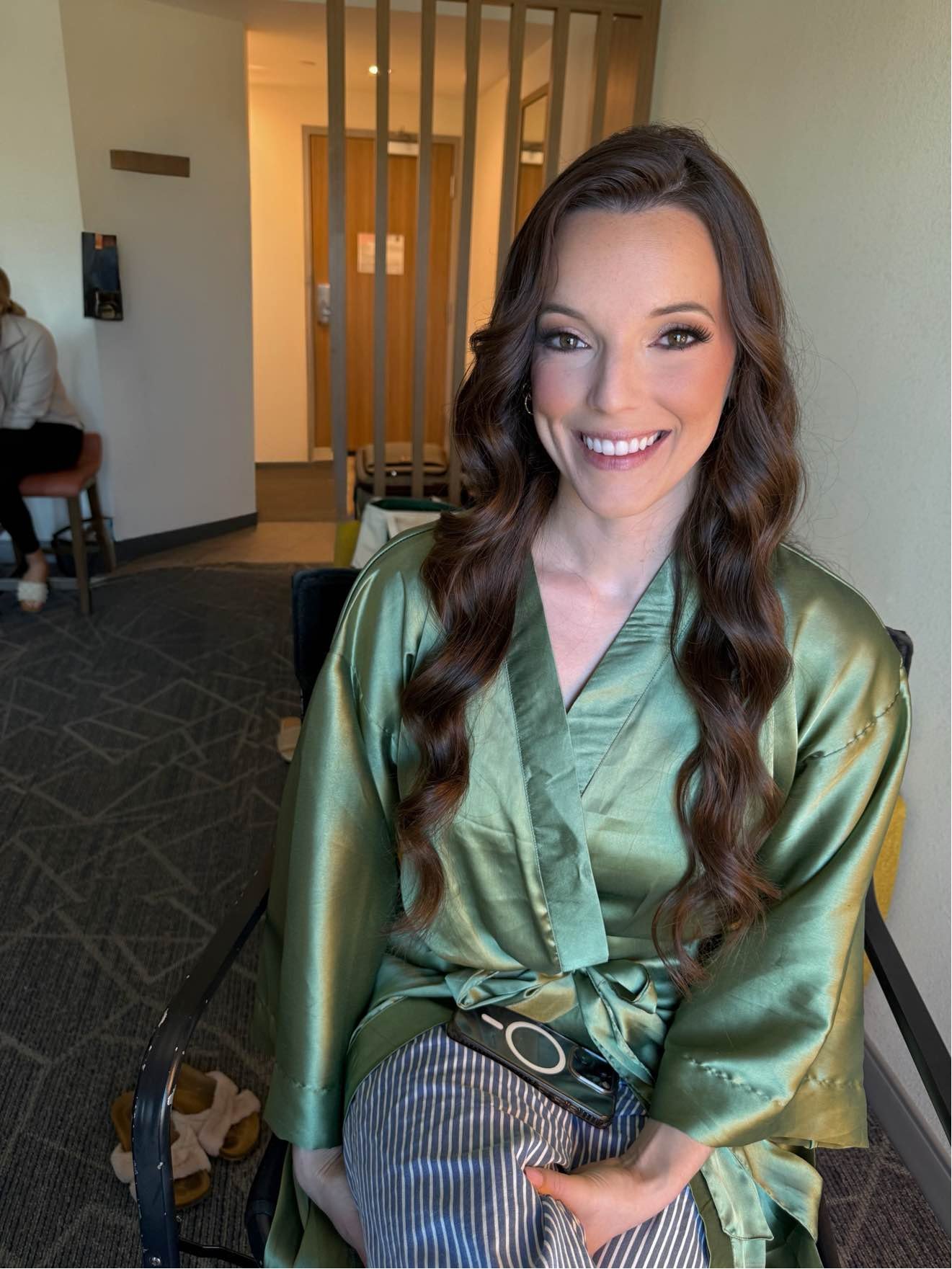 A woman with long wavy brown hair and makeup smiling at the camera, wearing a shiny green satin robe, seated indoors with wooden and beige background elements, and a person sitting on a chair in the background.