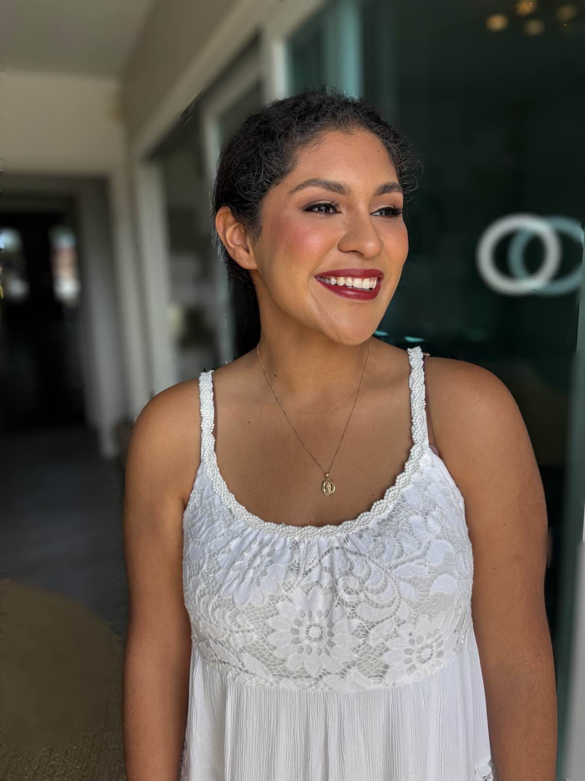 A woman with short curly hair smiling and wearing a white lace dress and a gold necklace, standing near glass doors.