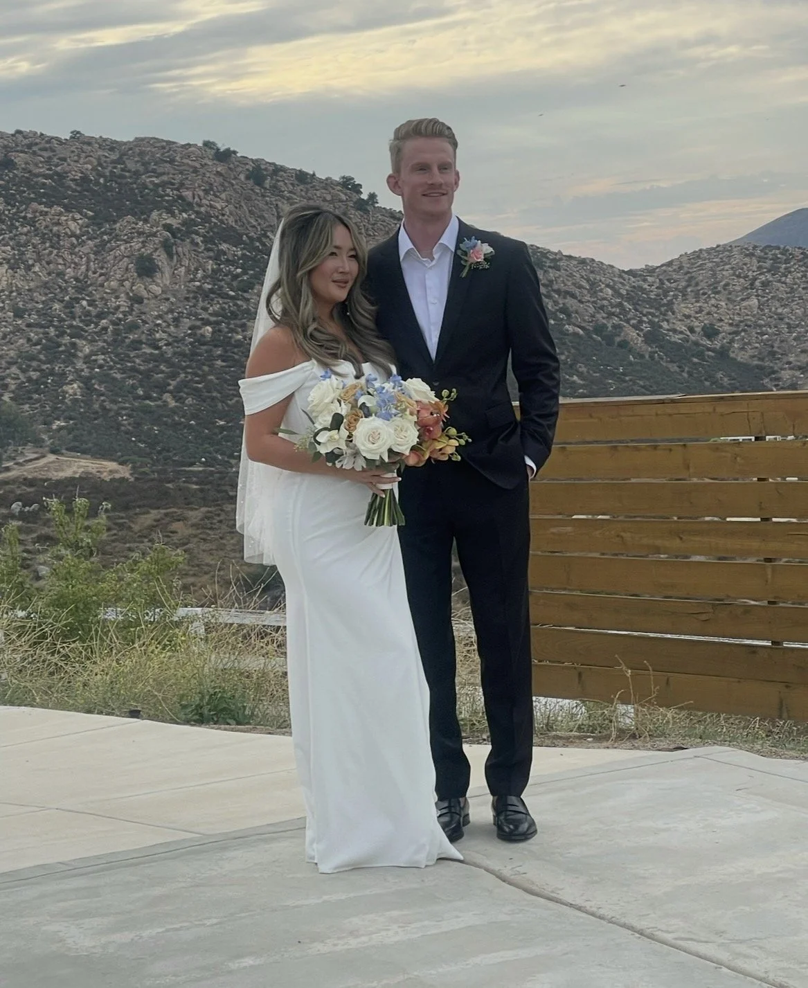 A bride and groom at their outdoor wedding, standing on a concrete surface with mountains in the background. The bride is holding a bouquet of flowers, wearing a white off-shoulder dress and a veil. The groom is dressed in a black suit with a white s