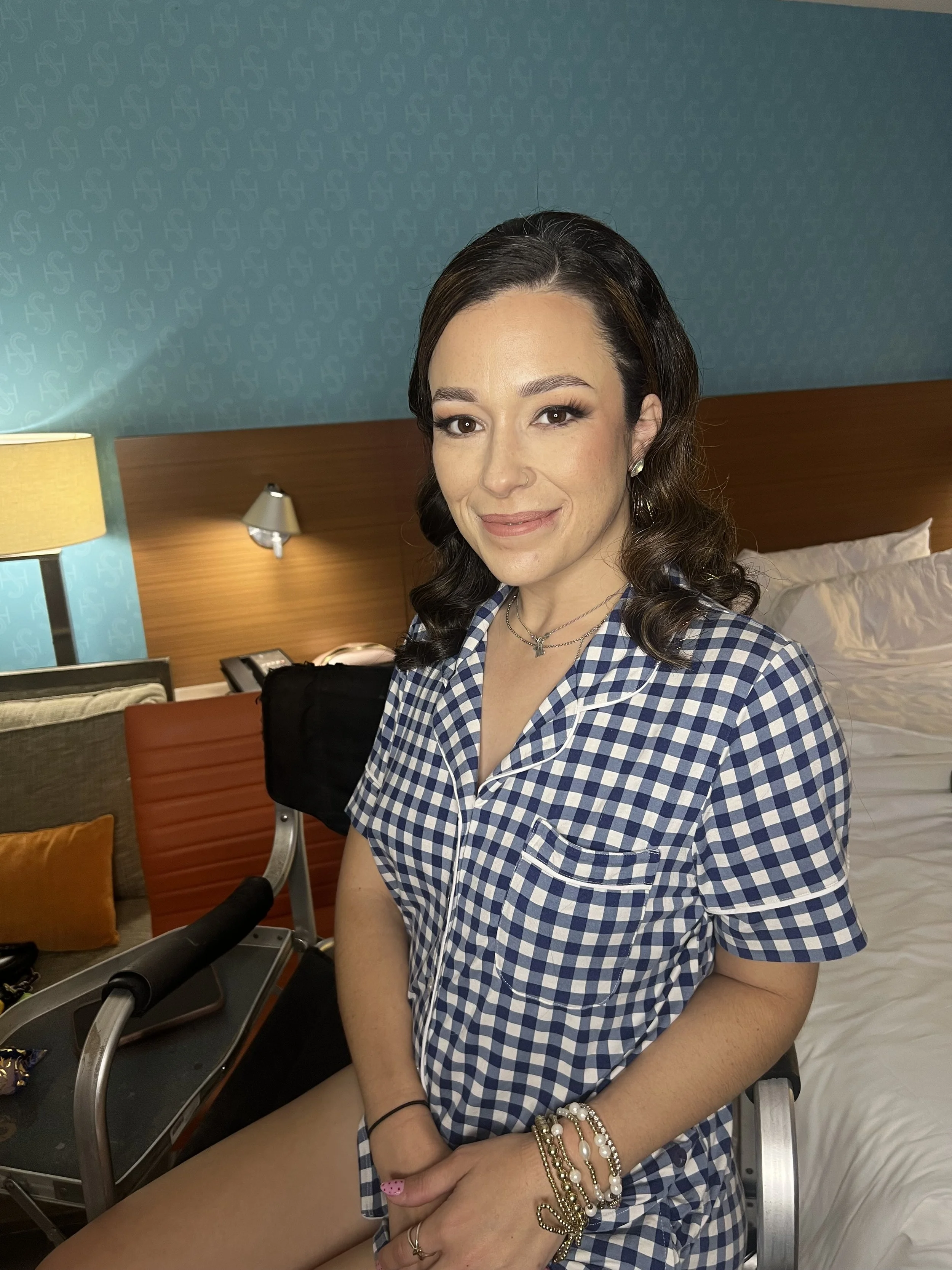 A young woman with dark hair and light makeup sitting on a bed in a hotel room, wearing a blue and white checkered shirt and multiple bracelets, smiling slightly, with her arm resting on her lap.