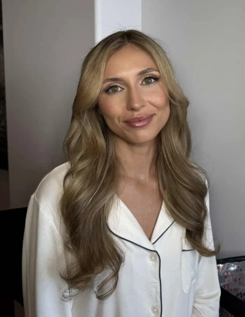 A woman with long, wavy blonde hair, wearing a white silk pajama top with black piping, smiling at the camera.
