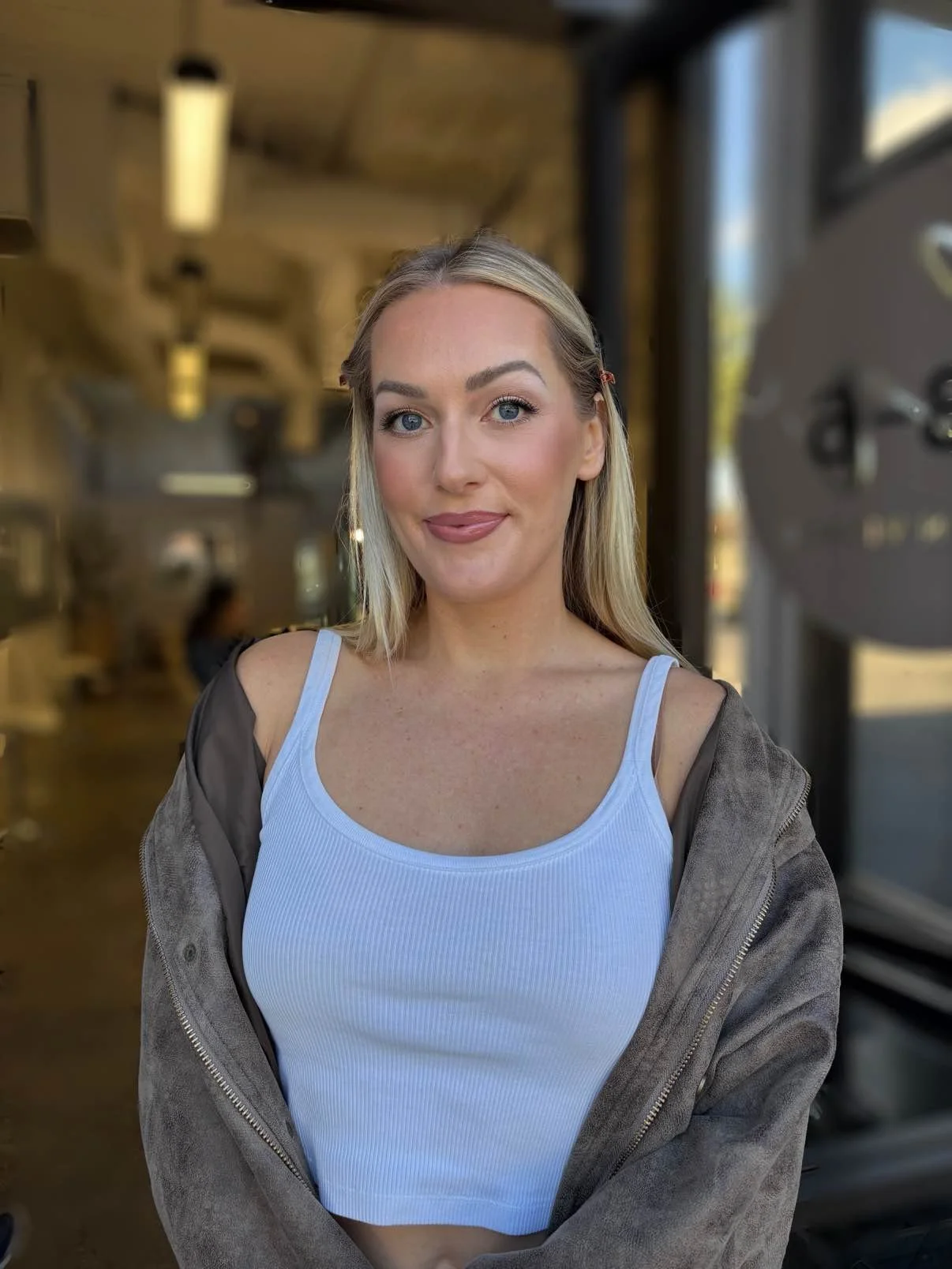 A young woman with long blonde hair and blue eyes, wearing a white tank top and a brown jacket, standing inside a modern cafe or restaurant with a blurred background.