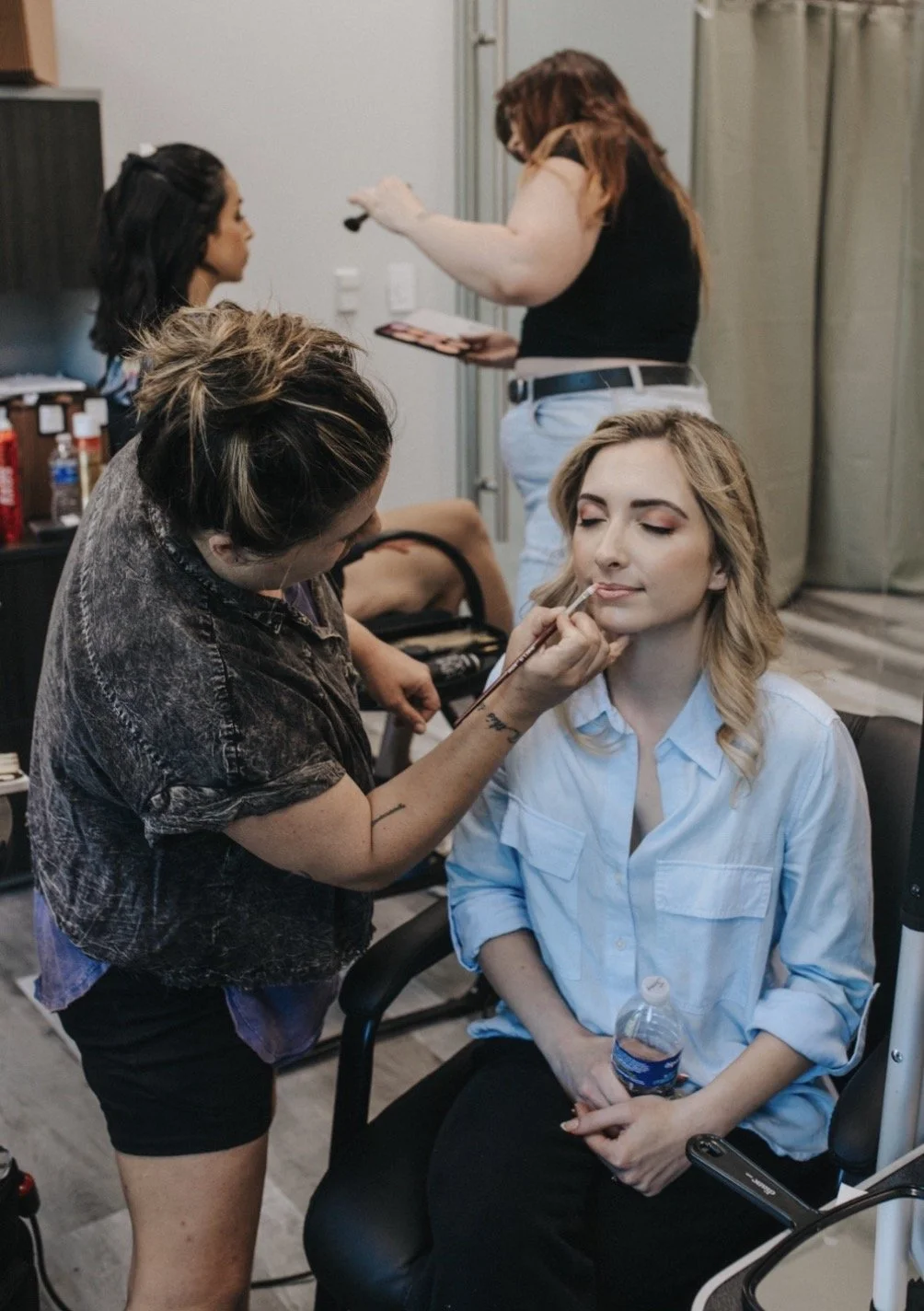 A woman in a white shirt sits with closed eyes while a makeup artist applies makeup on her face. Another woman is seen in the background, preparing makeup products.